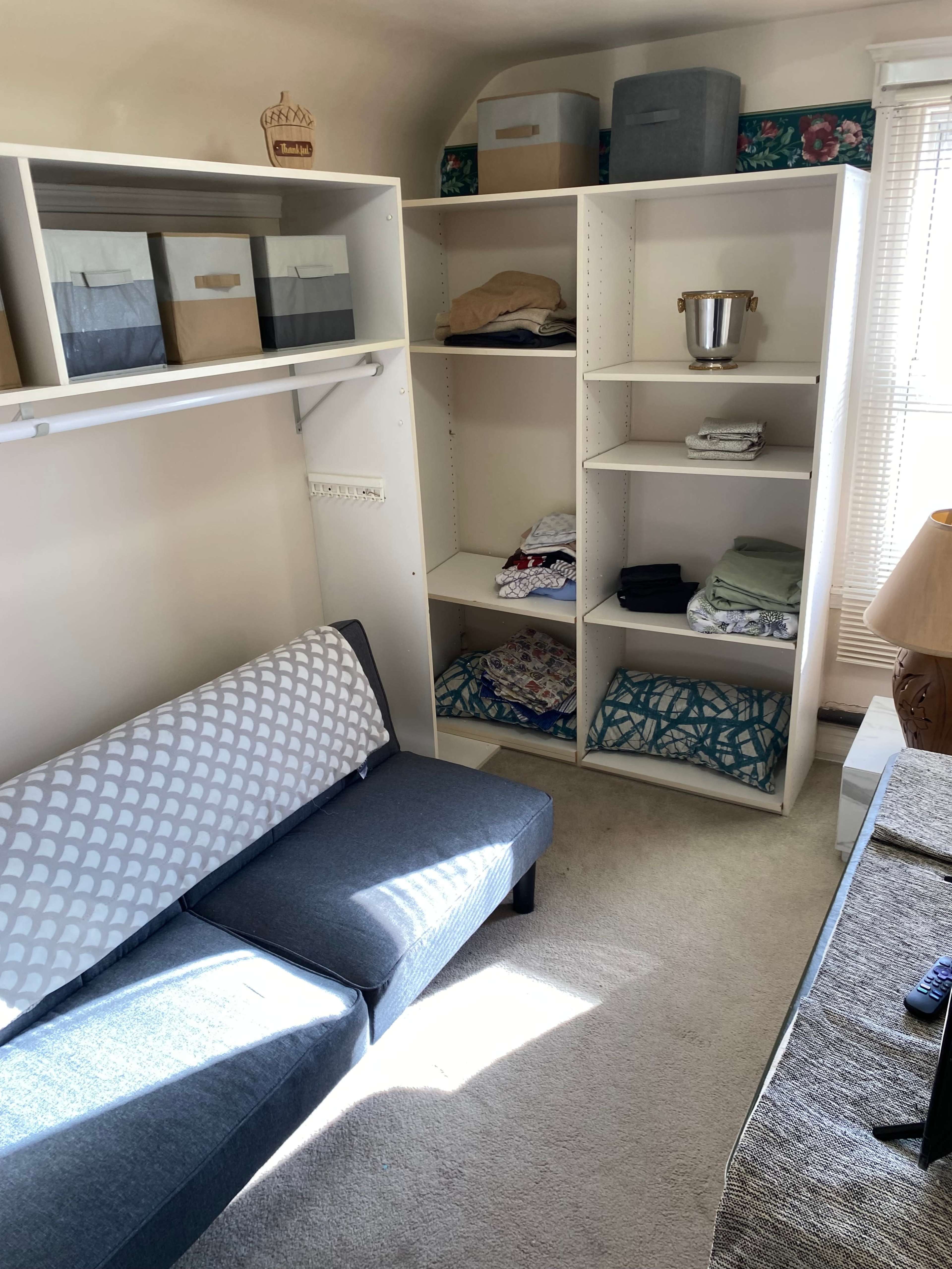 The image shows a neatly organized small room featuring a grey couch, a shelving unit filled with folded blankets and storage boxes, and a window letting in natural light.