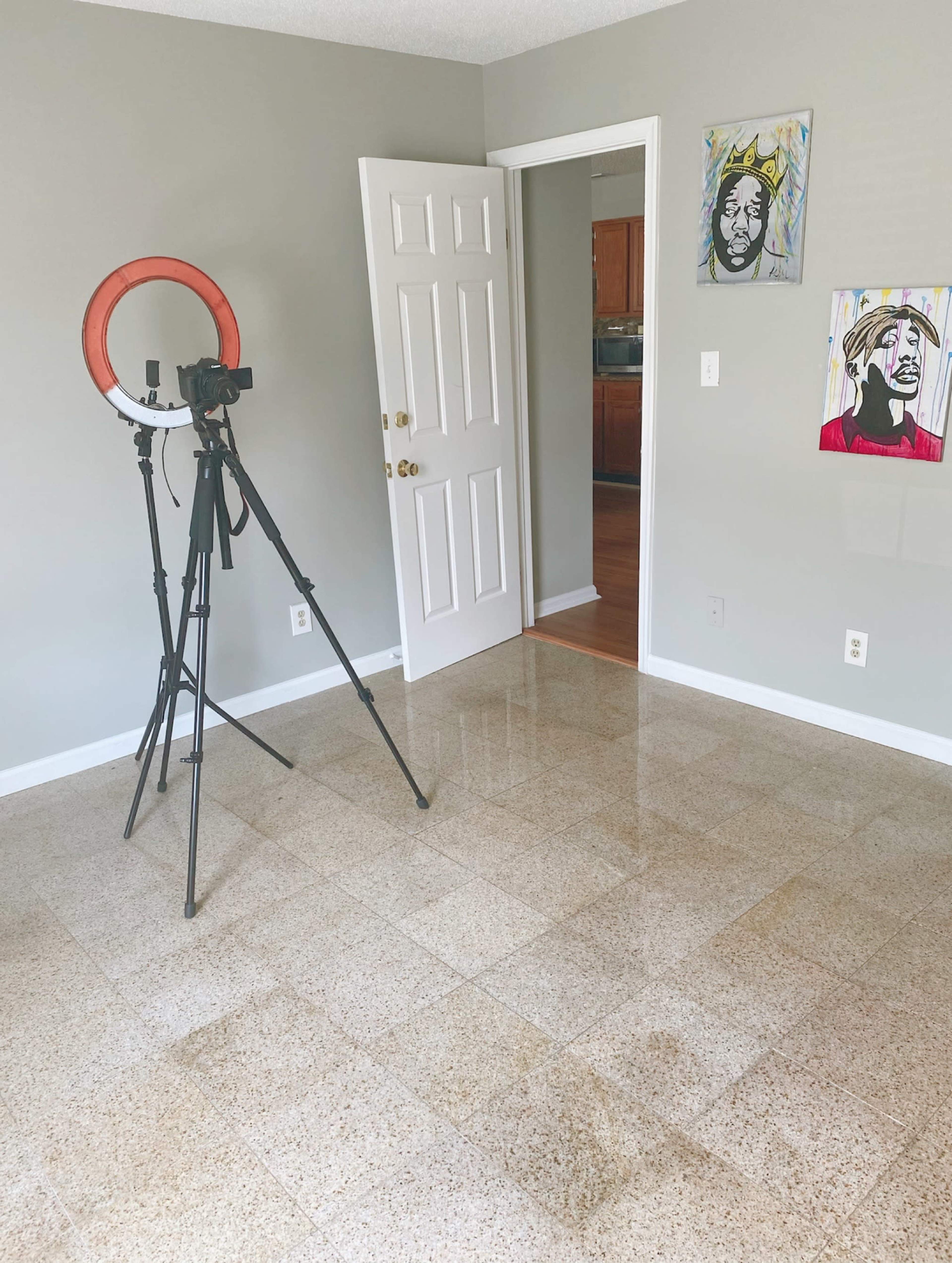 A room with a tripod and ring light set up, light gray walls, and two framed portraits hanging on the wall.