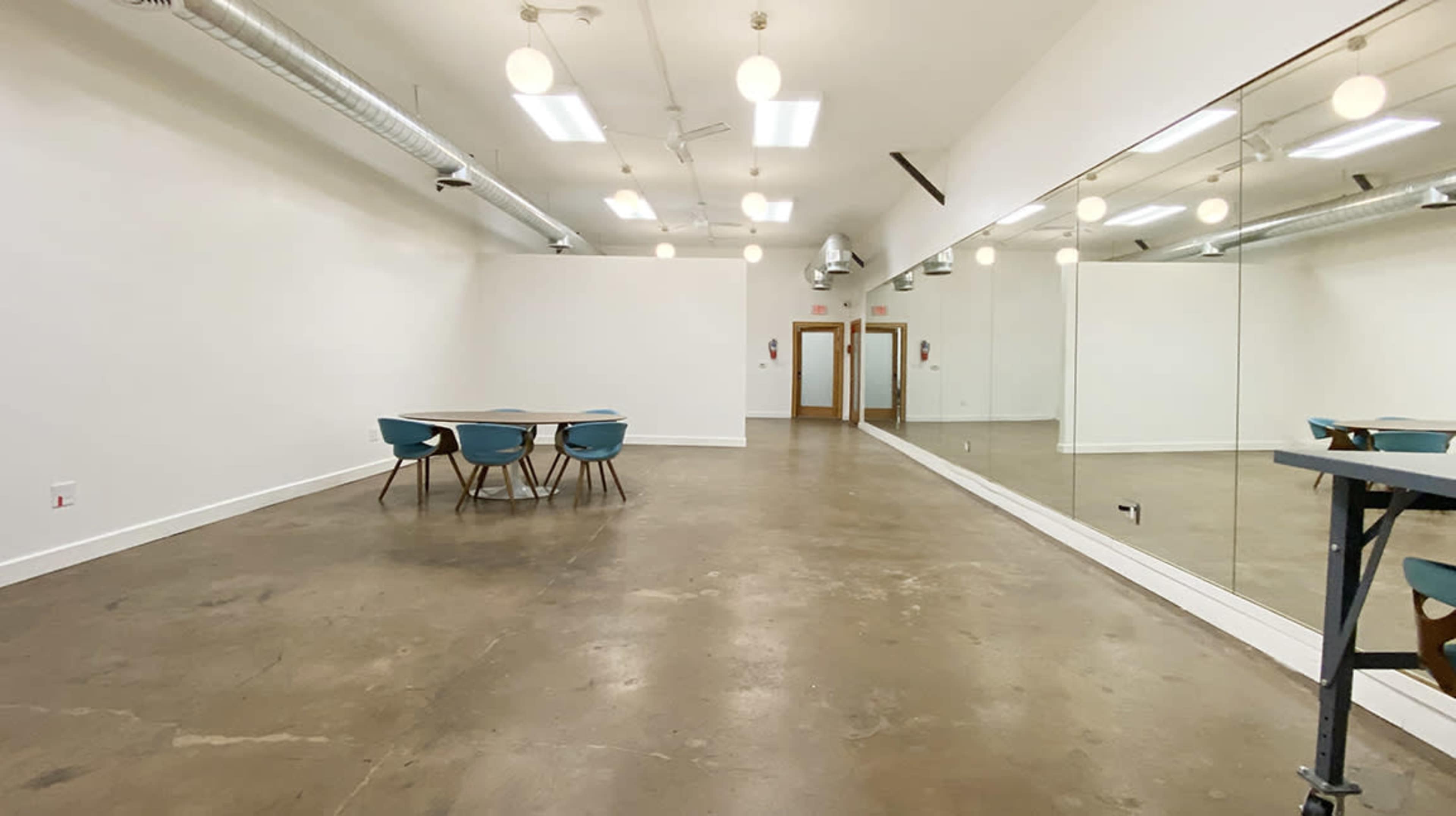 The image shows a spacious room with polished concrete flooring, a set of teal chairs around a table, and a large mirror along one wall.