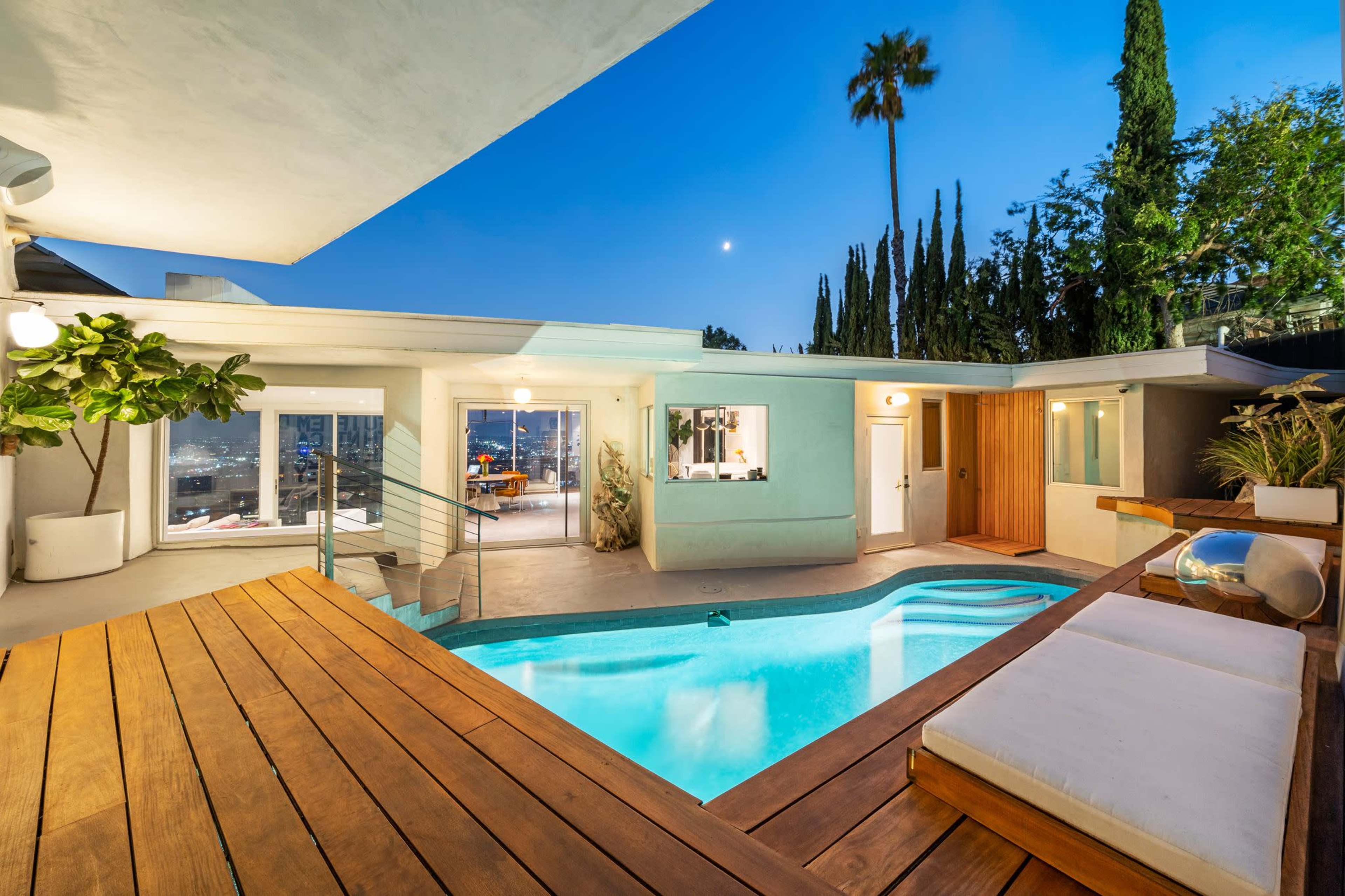 The image shows a modern patio with a swimming pool, surrounded by palm trees and illuminated under a twilight sky.
