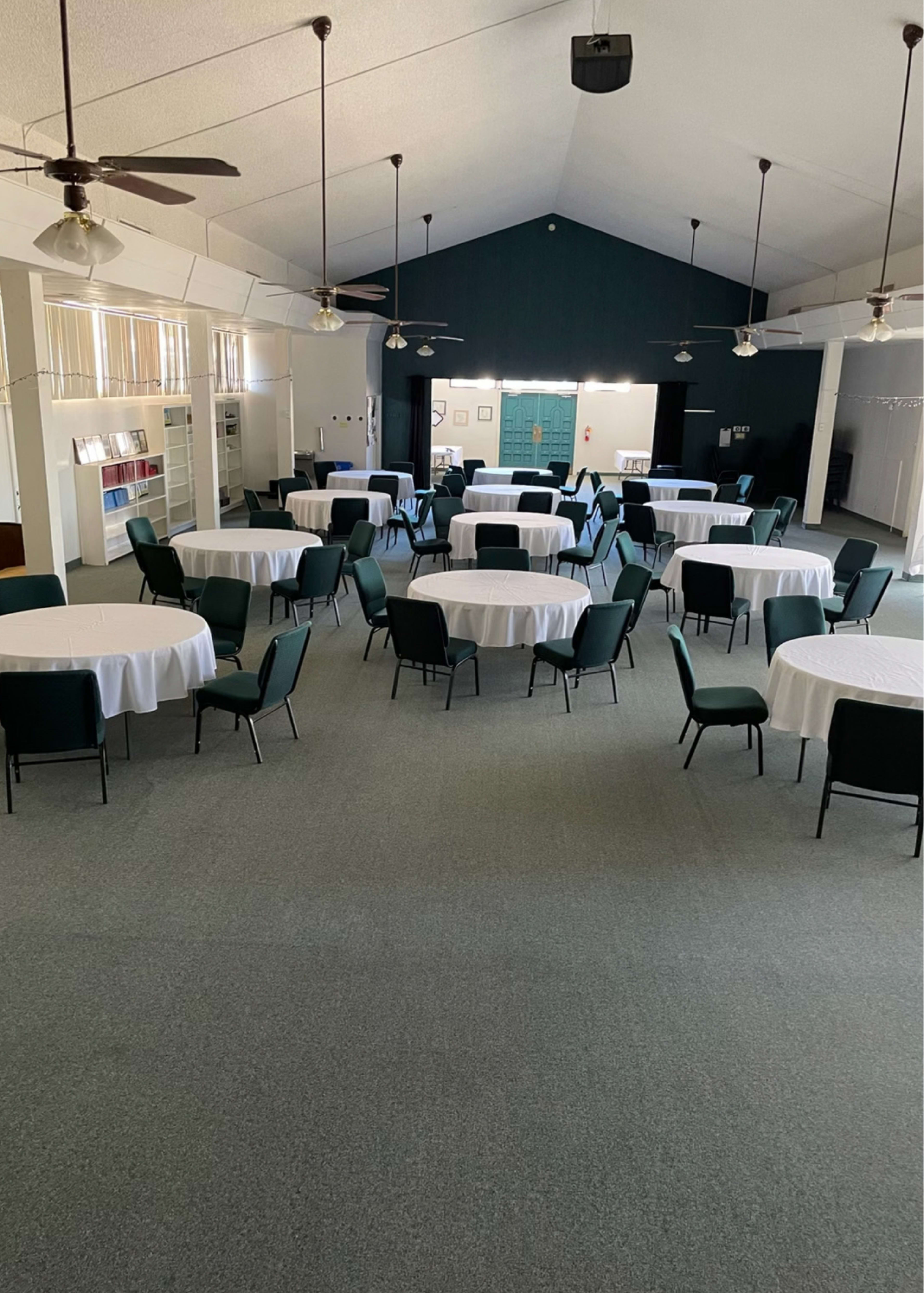 The image shows a spacious room with several round tables covered in white tablecloths, surrounded by green chairs, under a high ceiling with ceiling fans.