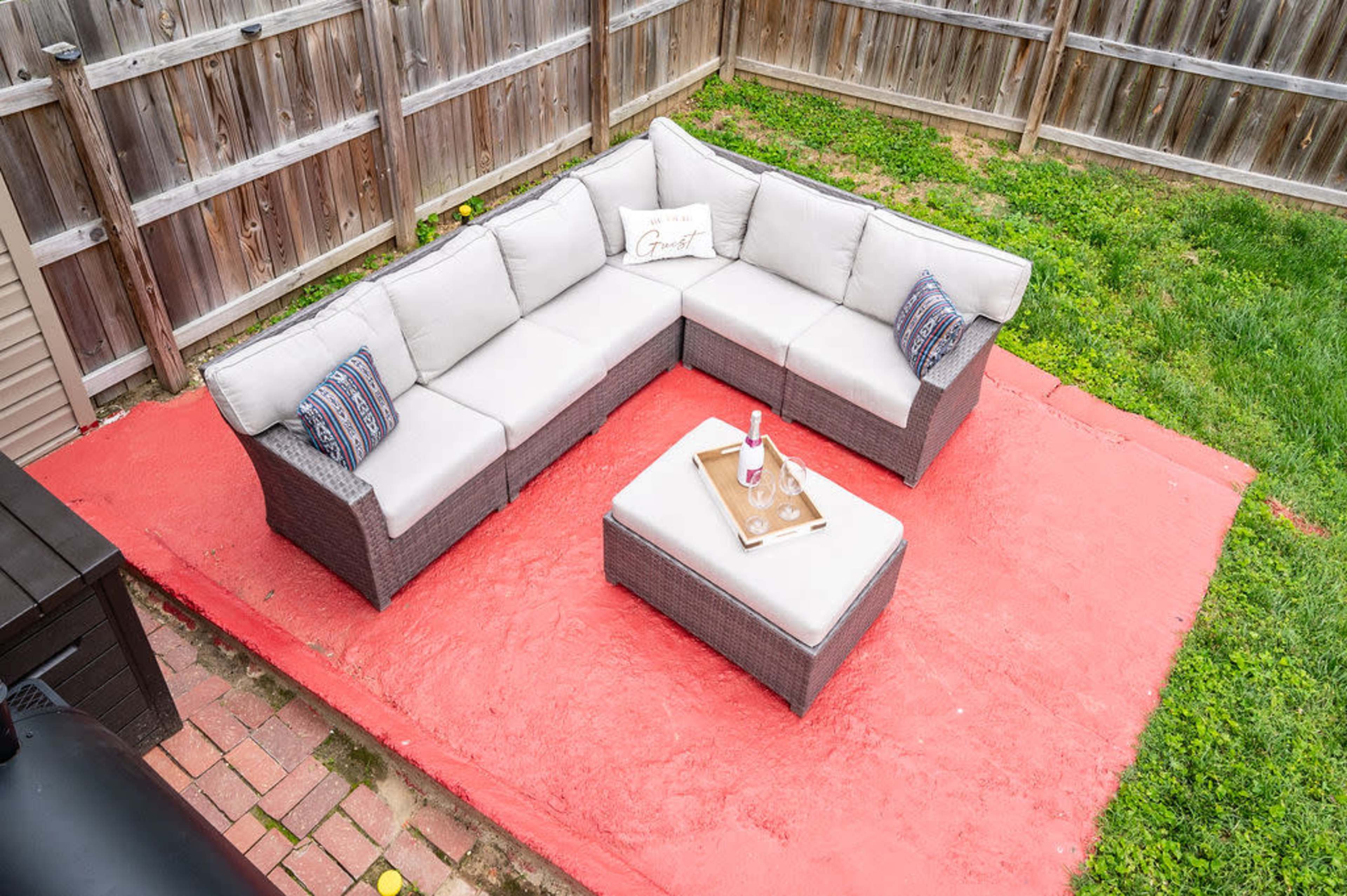 A sectional outdoor sofa with a coffee table is placed on a red patio in a fenced backyard.