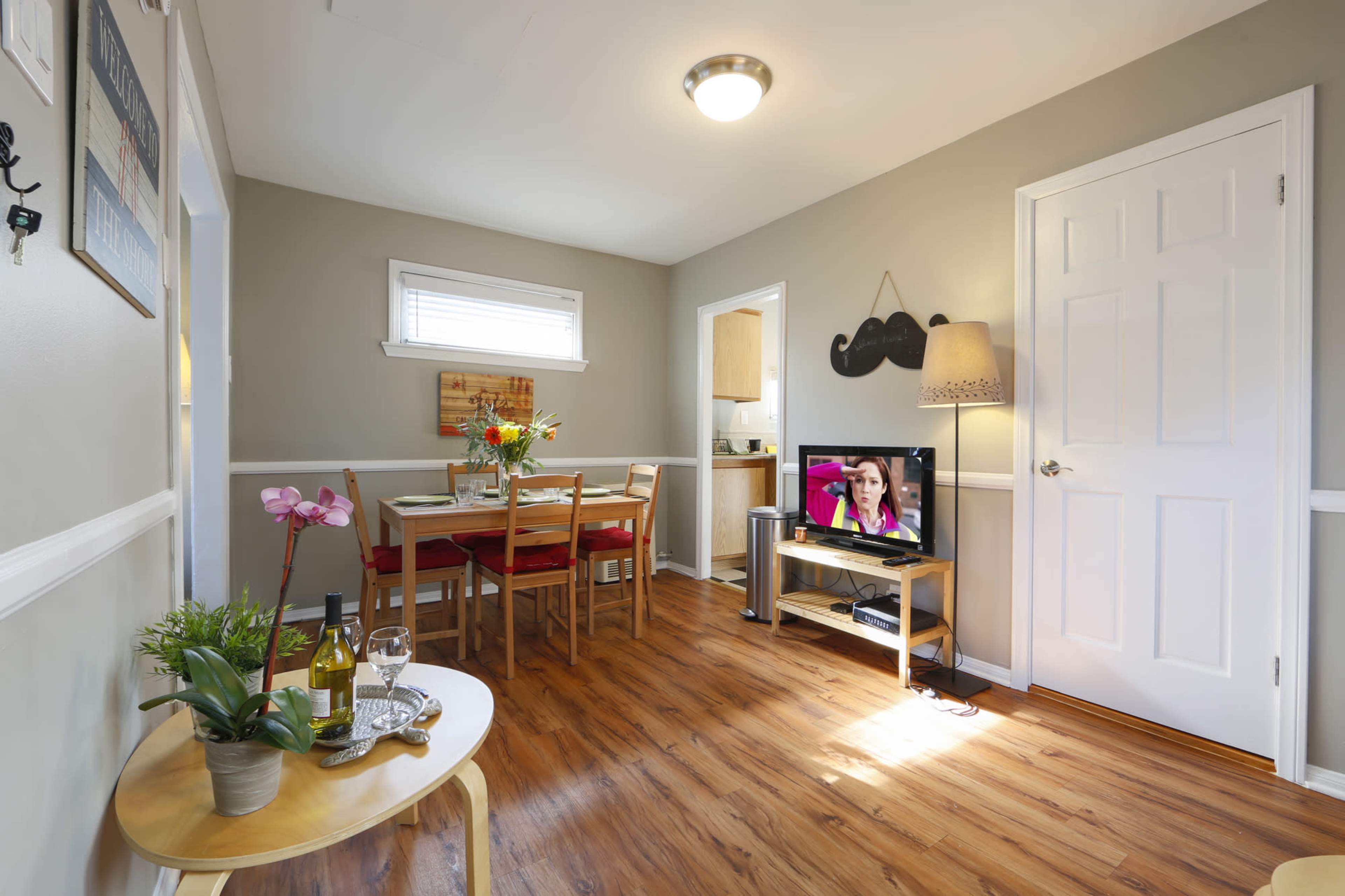 A cozy living space features a dining area with a table, chairs, and a television displaying a program, surrounded by light-colored walls and wooden flooring.