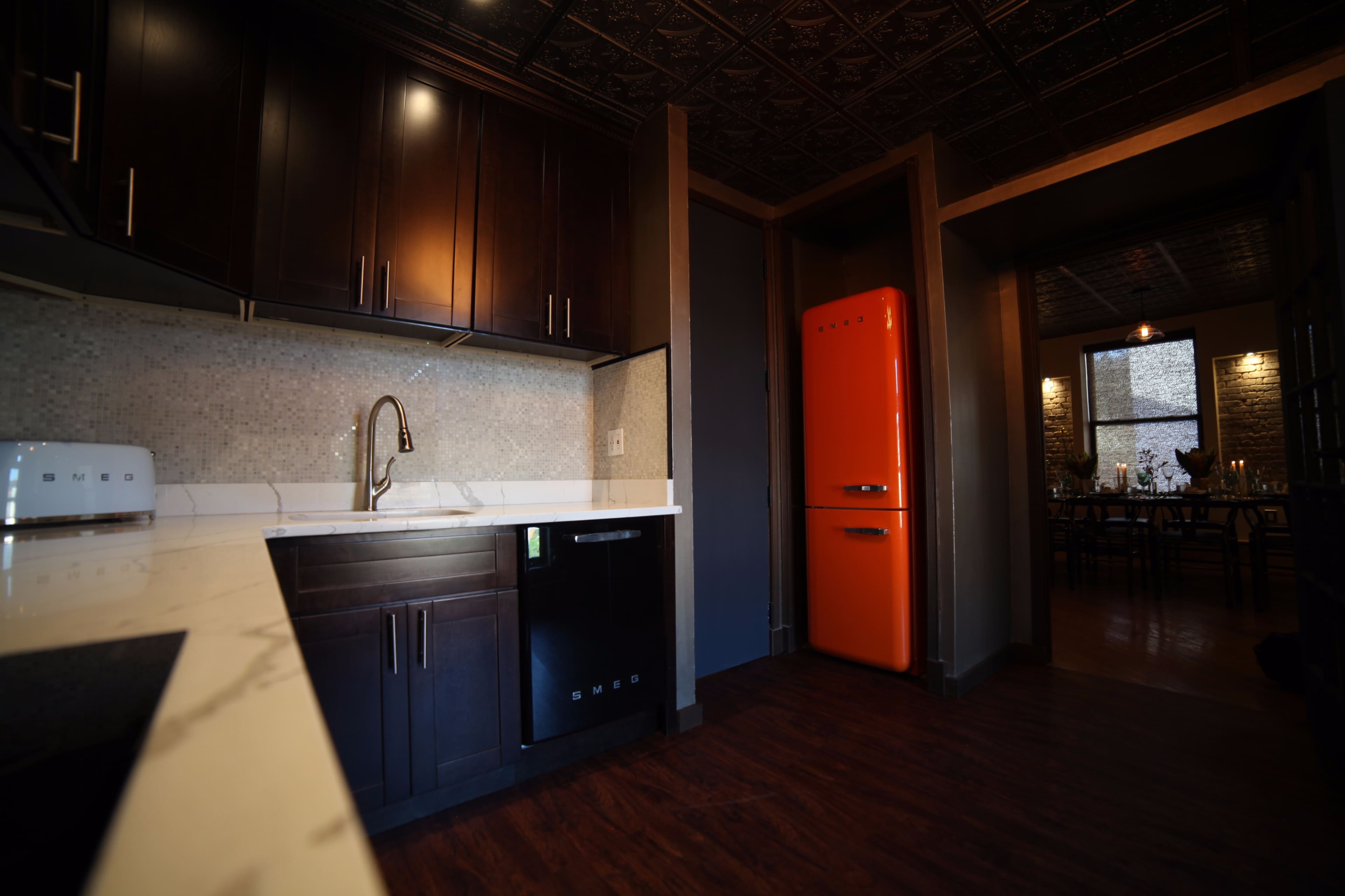 A modern kitchen featuring dark cabinetry, a white countertop, and an orange refrigerator positioned near a doorway leading to a dining area.