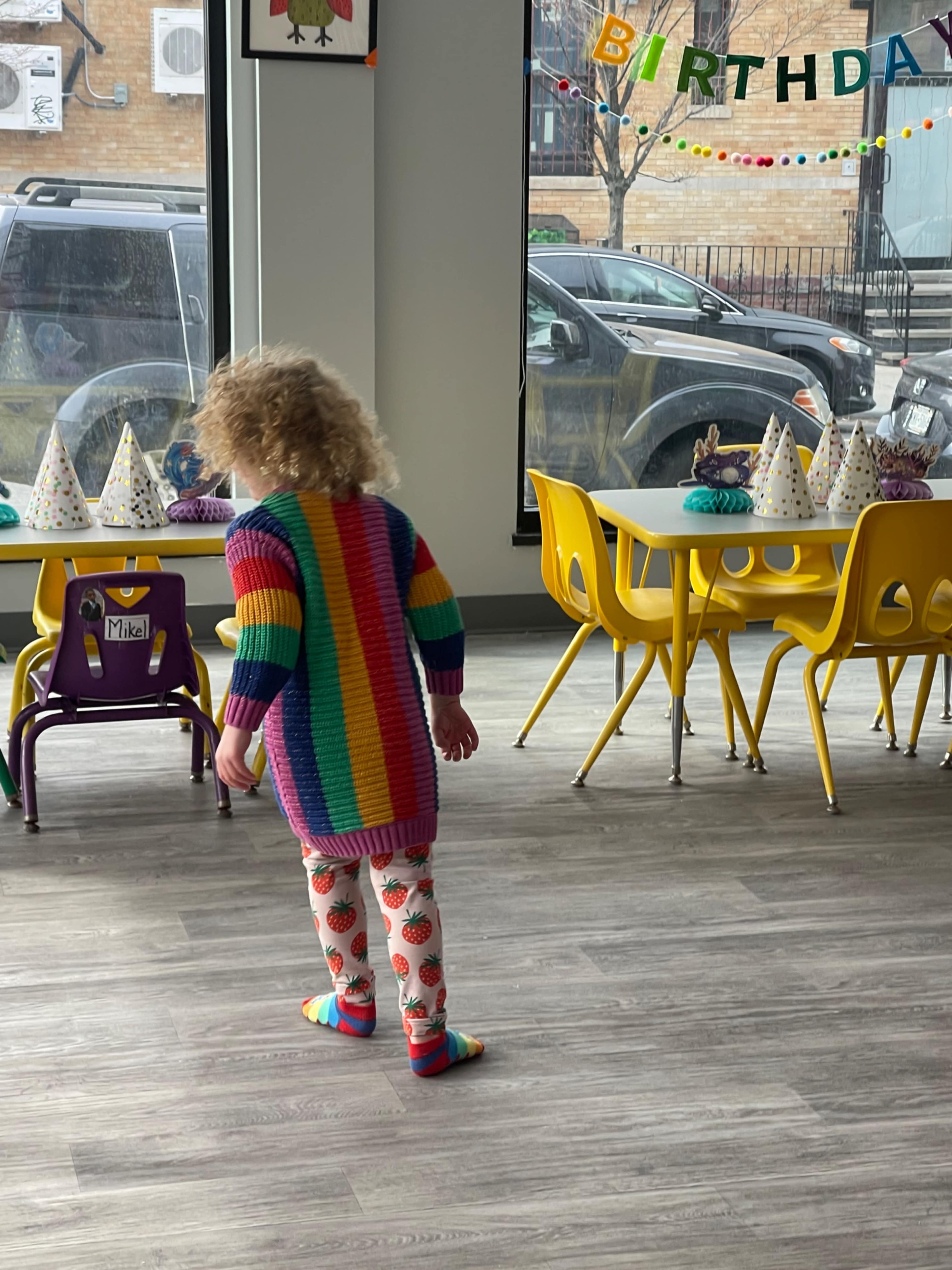 A child wearing a colorful sweater and patterned leggings walks across a room decorated for a birthday party, with yellow chairs and tables in the background.