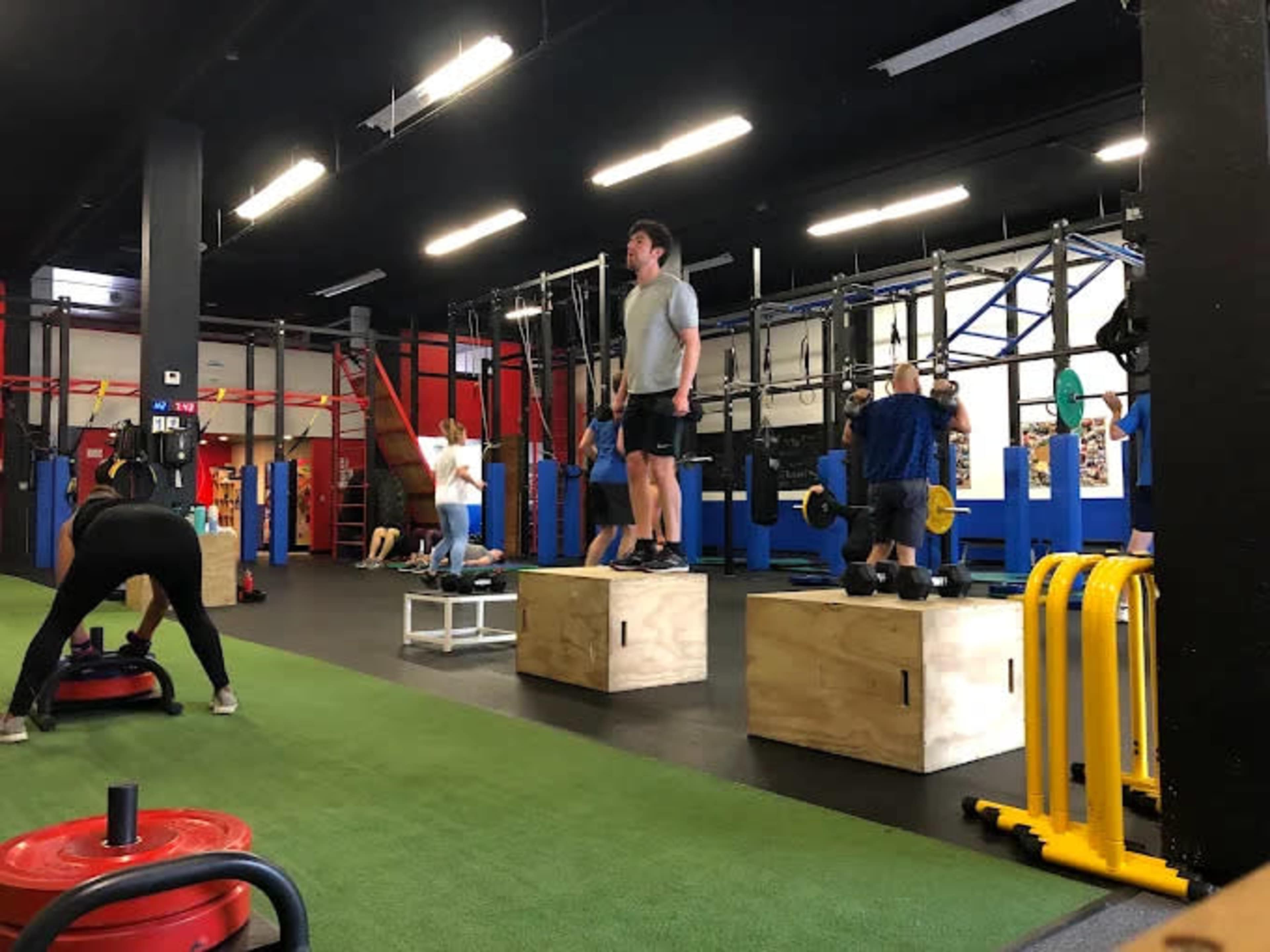 The image shows a gym setting with individuals performing various exercises on wooden boxes and using other fitness equipment.