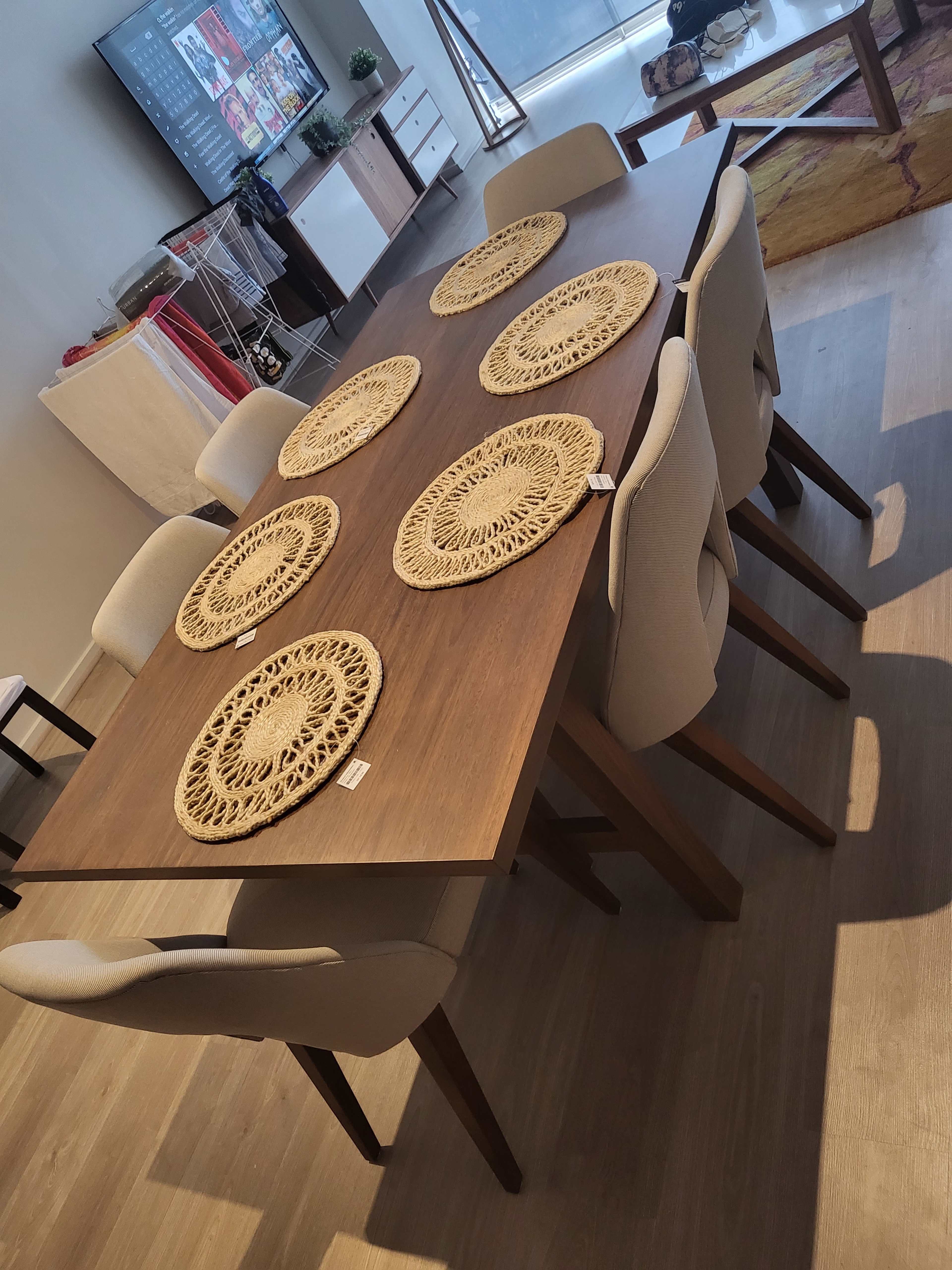 A wooden dining table with eight placemats is set in a room with modern furniture and a large window.