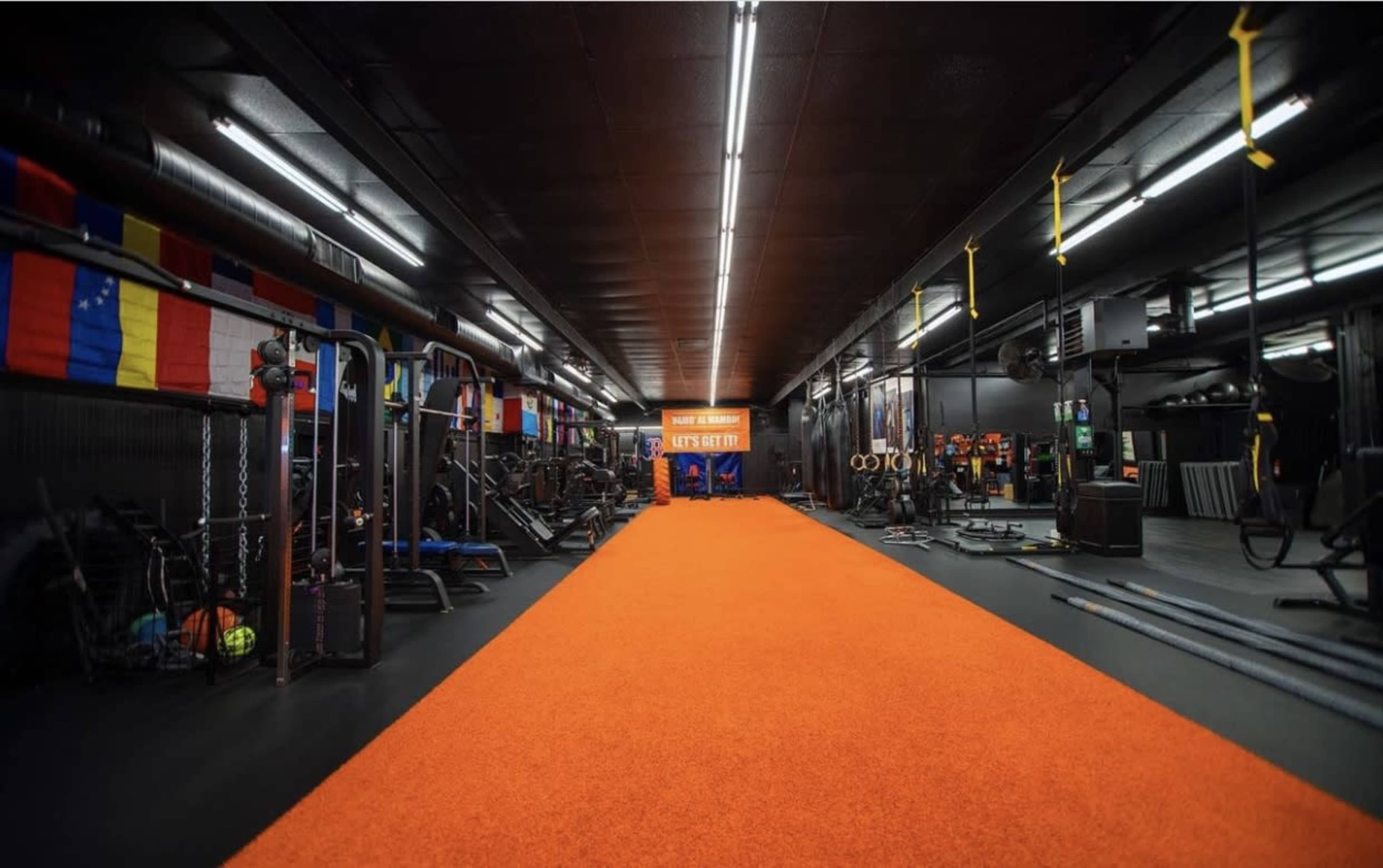 A spacious gym interior with exercise equipment on both sides and a bright orange carpet leading to an entrance in the distance.