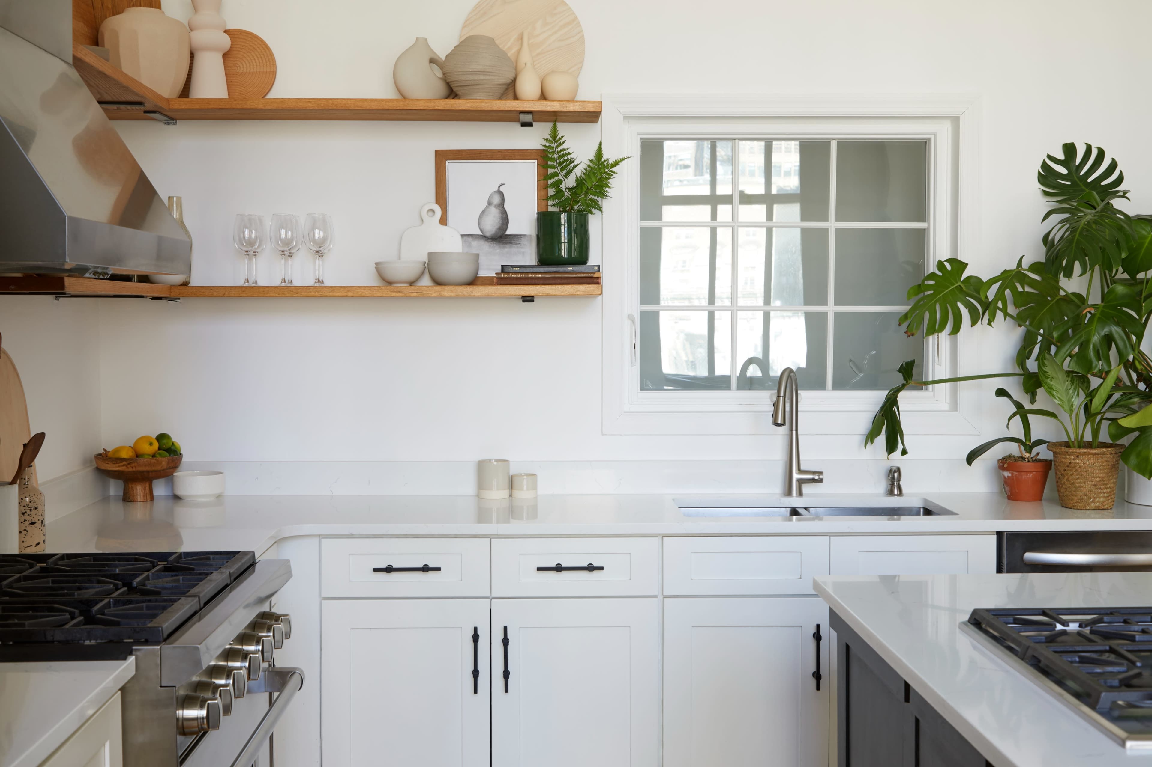 A modern kitchen features white cabinetry, open wooden shelves with decorative items, a stainless steel sink, and a large window.