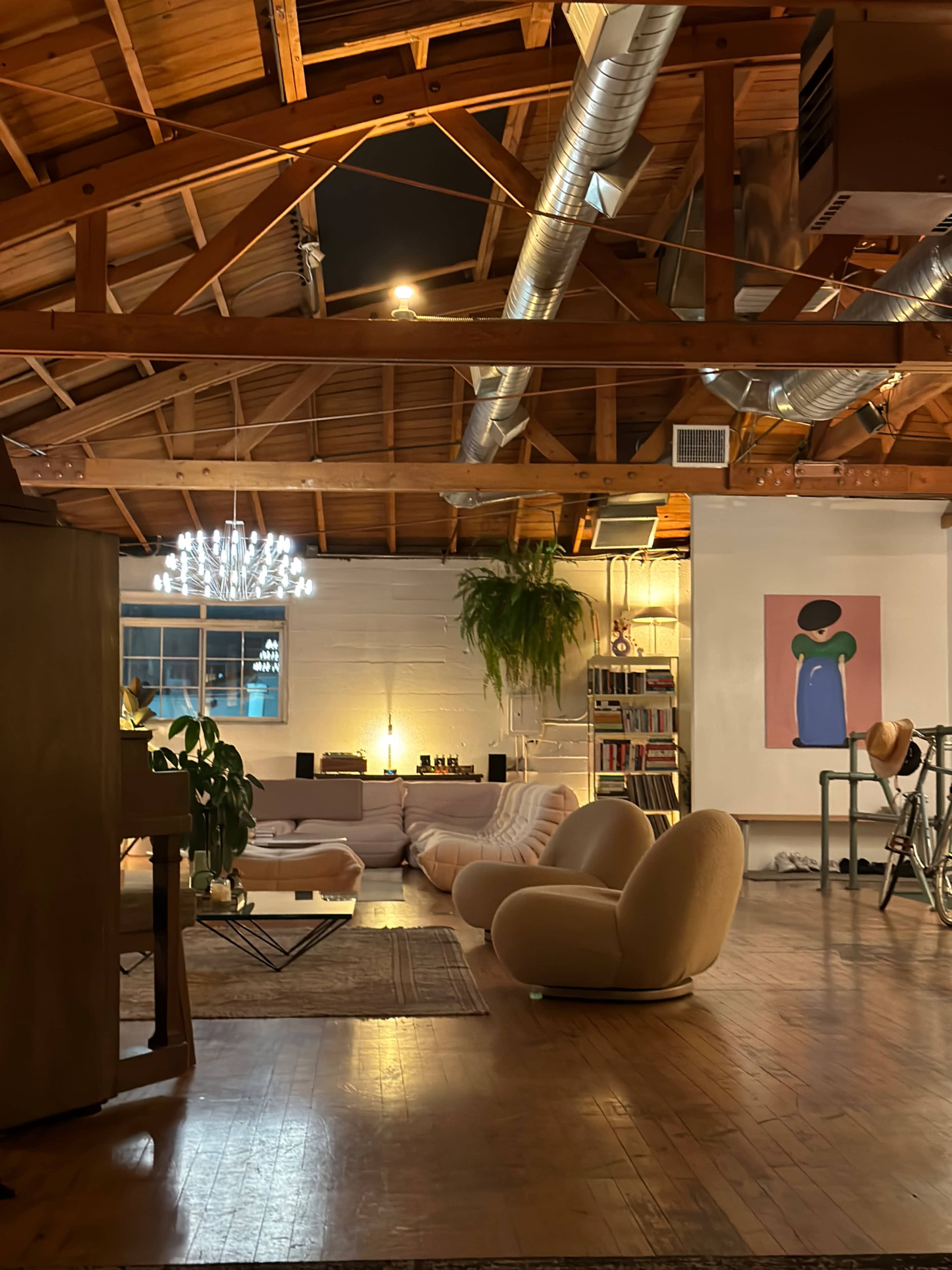 A spacious loft with wooden beams, a large sectional sofa, a round coffee table, and a colorful artwork on the wall, illuminated by soft lighting.