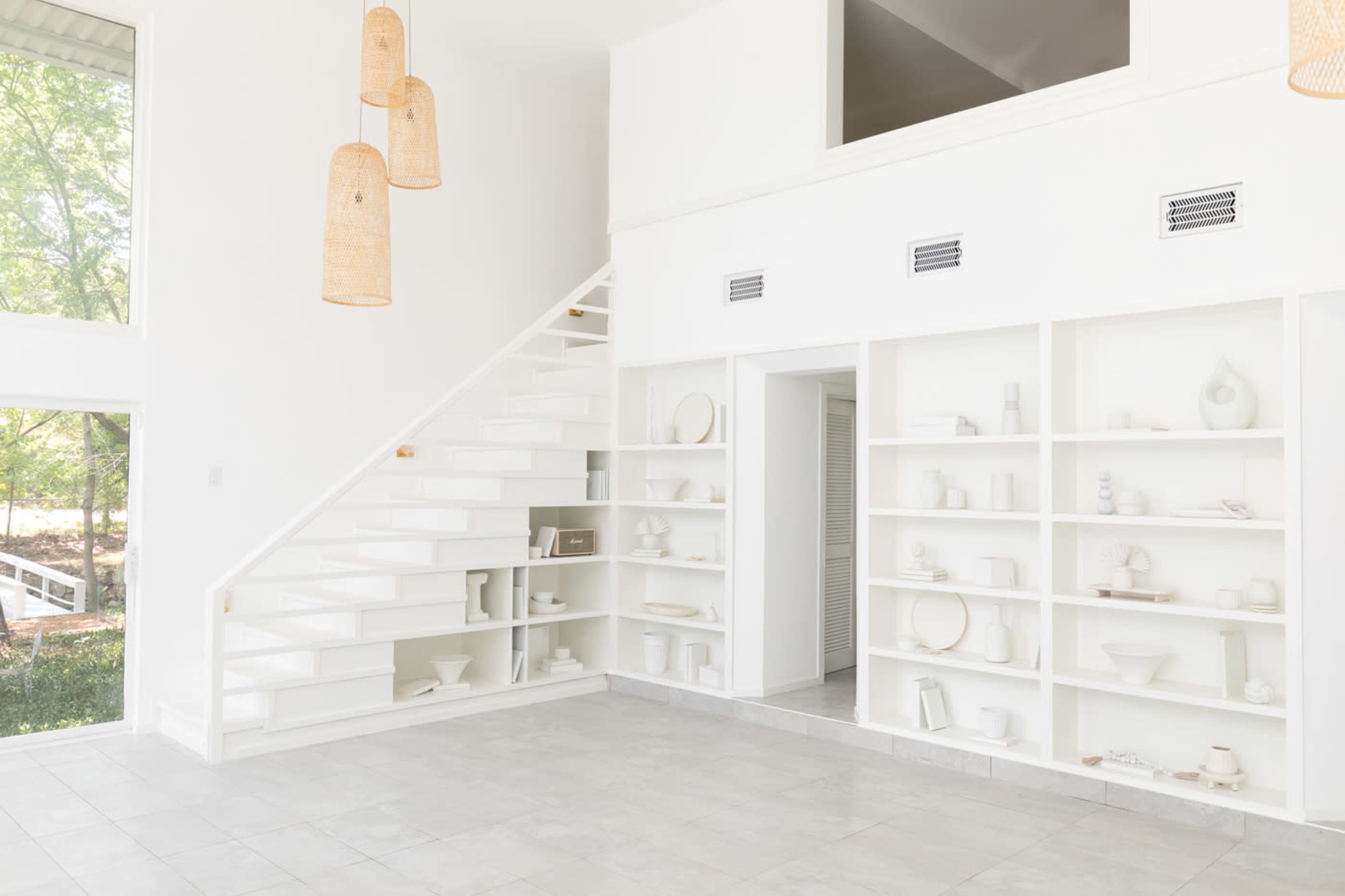 A bright, minimalist living space with white walls, a staircase, and built-in shelves displaying various white objects.