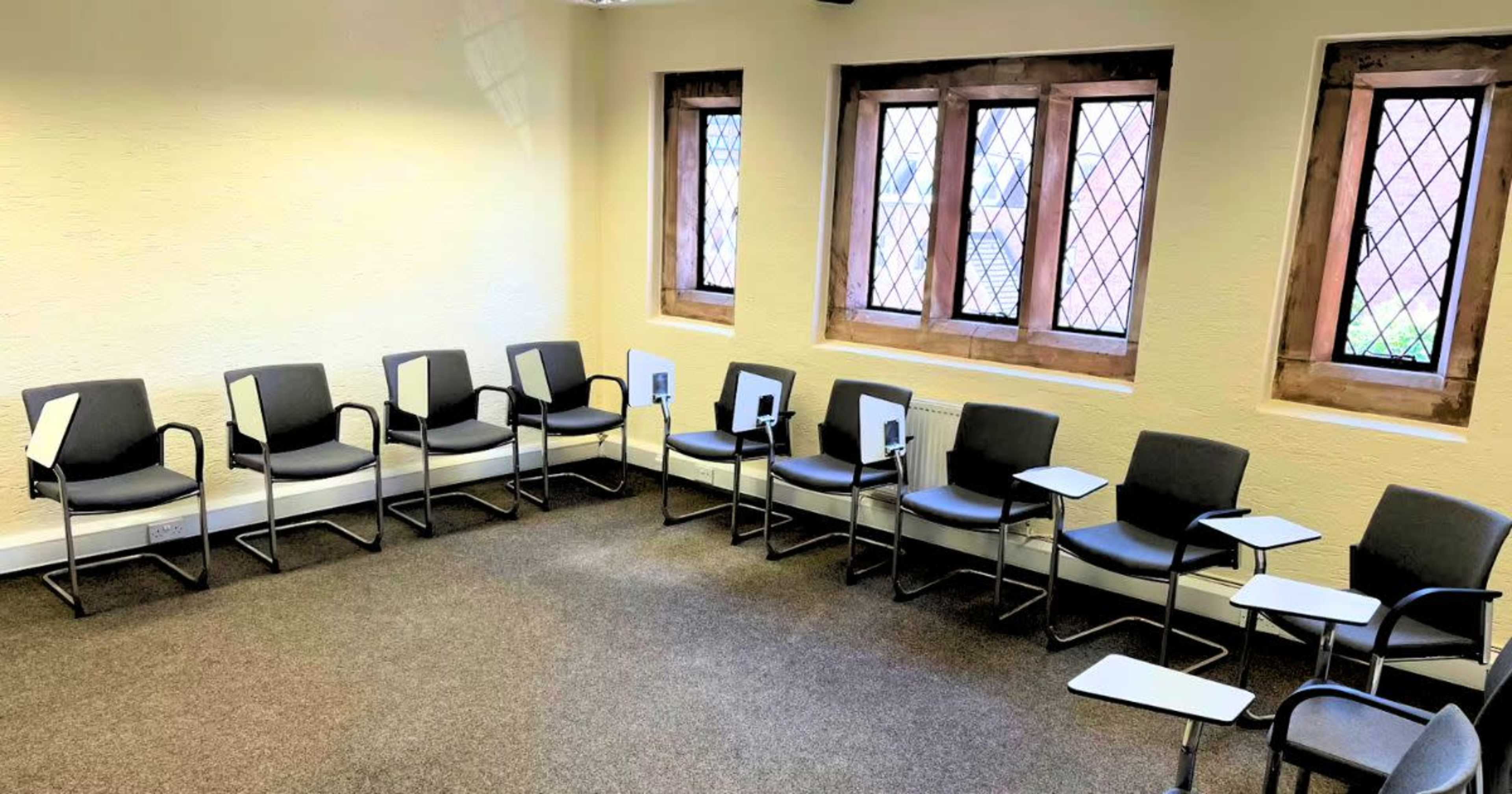 The image shows a classroom with a circular arrangement of gray chairs, each equipped with a small writing surface, next to windows with diamond-patterned panes.