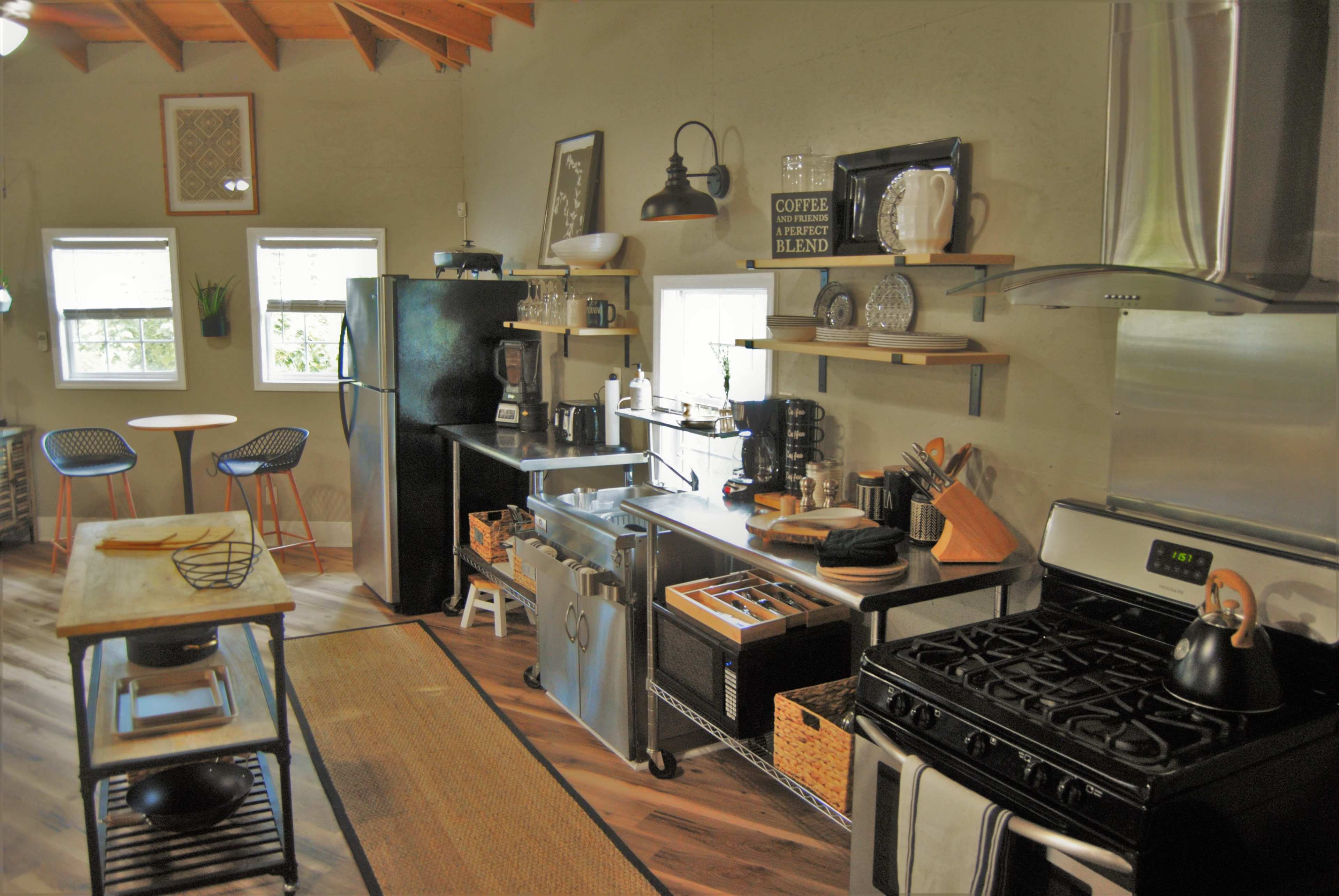 A modern kitchen featuring a black refrigerator, stainless steel appliances, wooden shelves with dishware, and a dining table with two chairs.