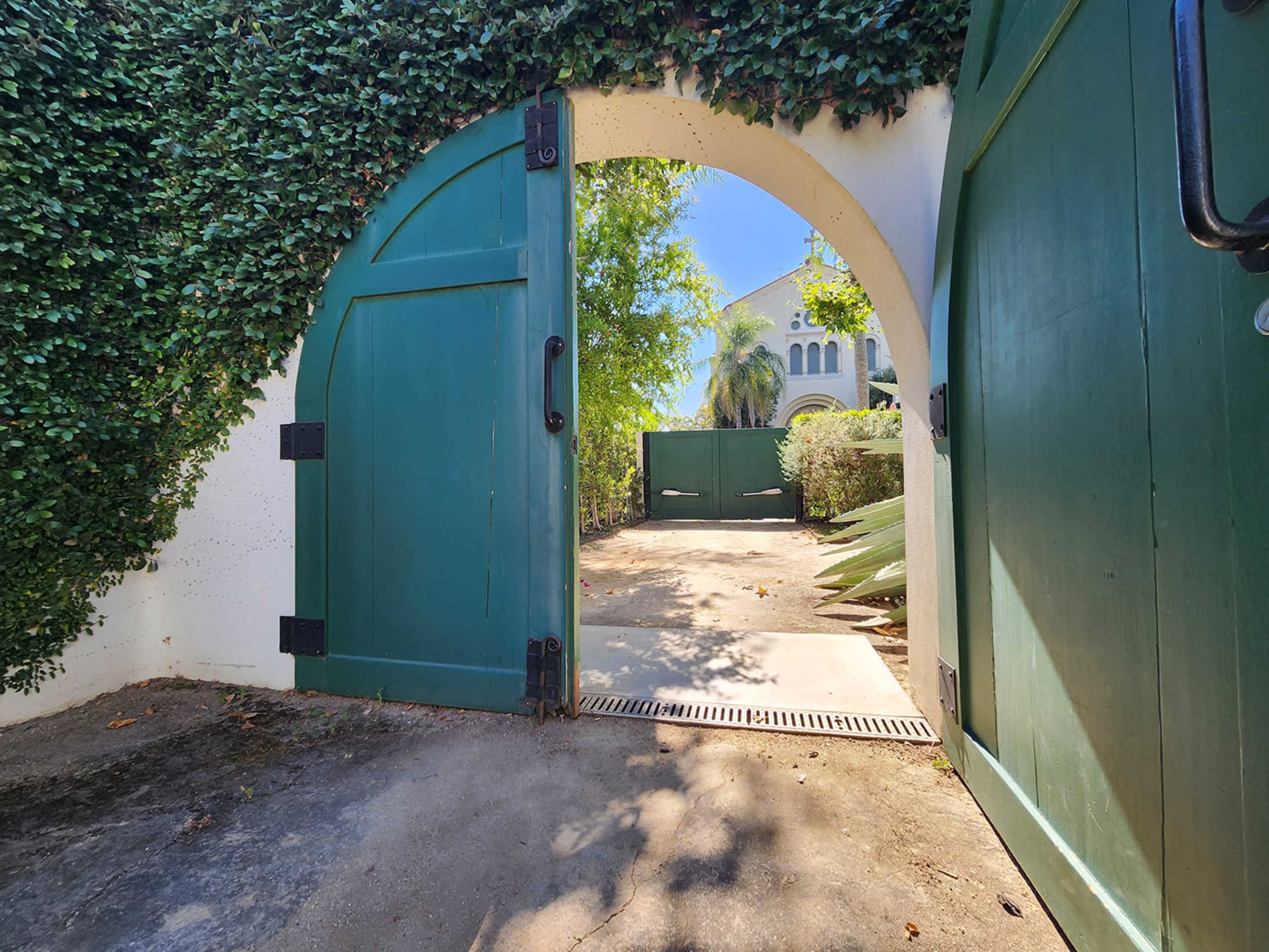 An arched green gate opens to a sunny pathway surrounded by lush foliage and a white wall.