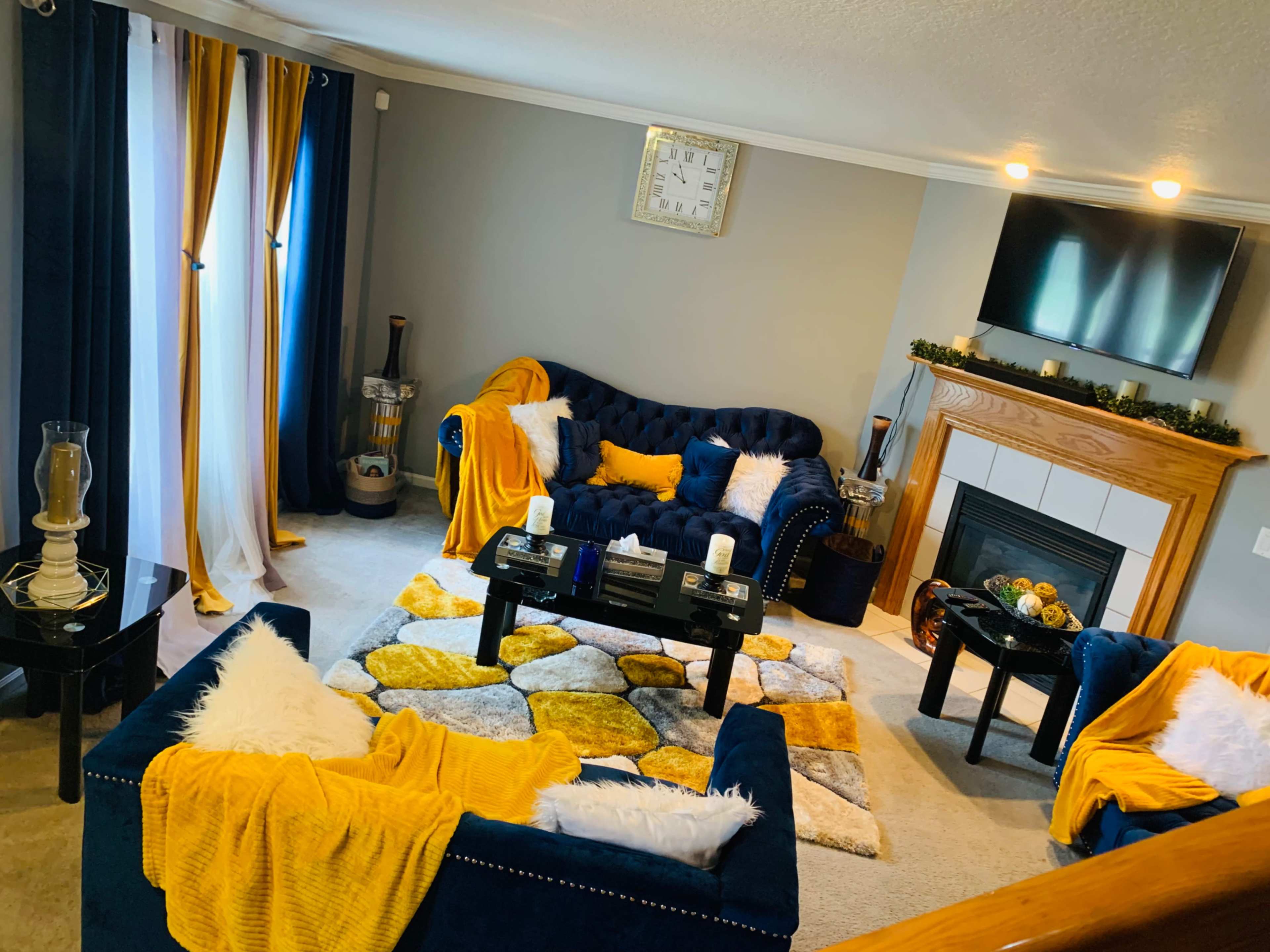 The living room features a navy blue sofa set, a fireplace with a wooden mantel, and a patterned area rug with yellow and gray tones.