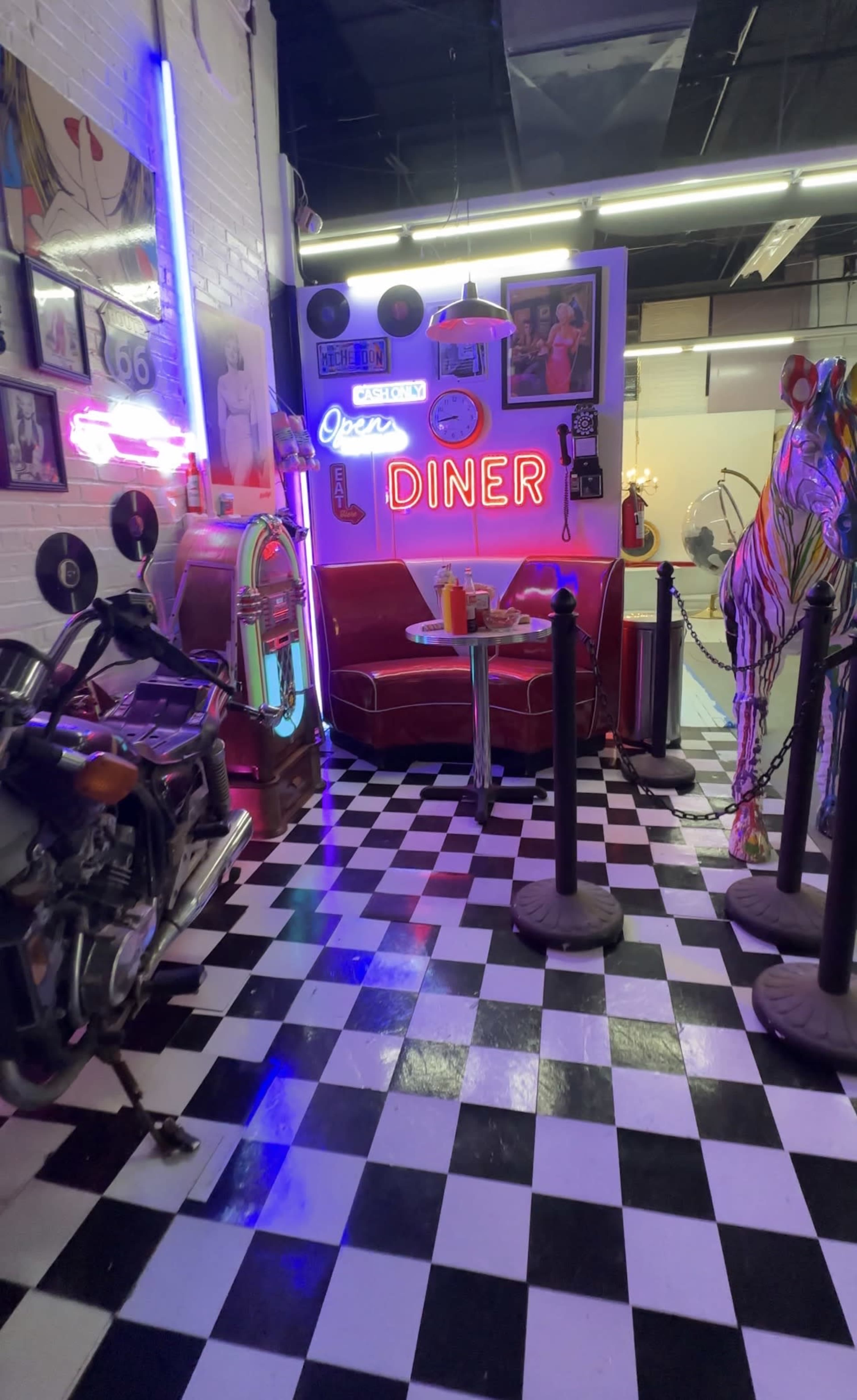 The image shows a vibrant diner interior with a checkered floor, neon signs, a retro jukebox, and colorful decor, including a horse sculpture and motorcycle.