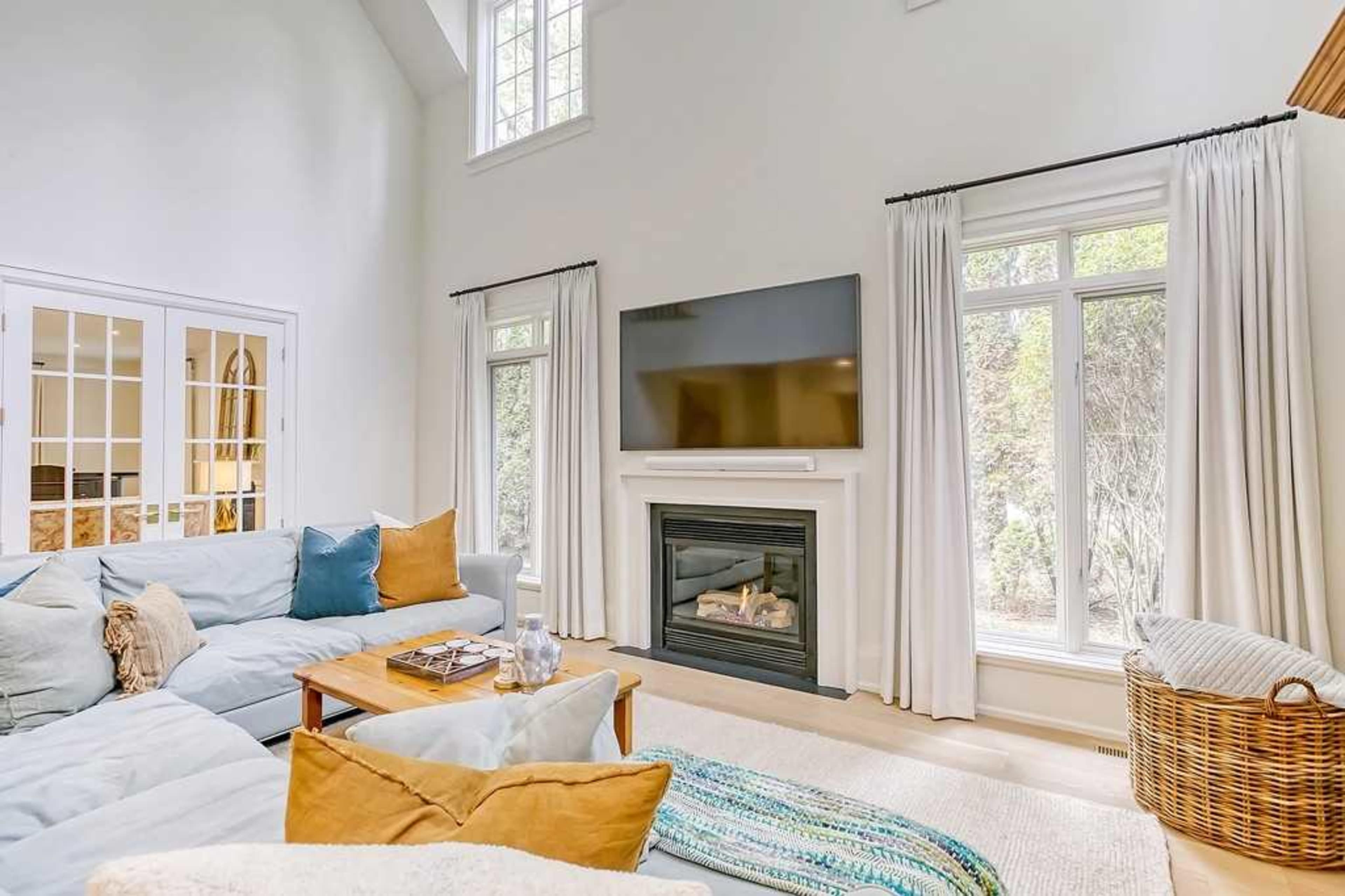 The living room features a large sectional sofa, a contemporary fireplace, and a mounted television with large windows allowing natural light to enter.