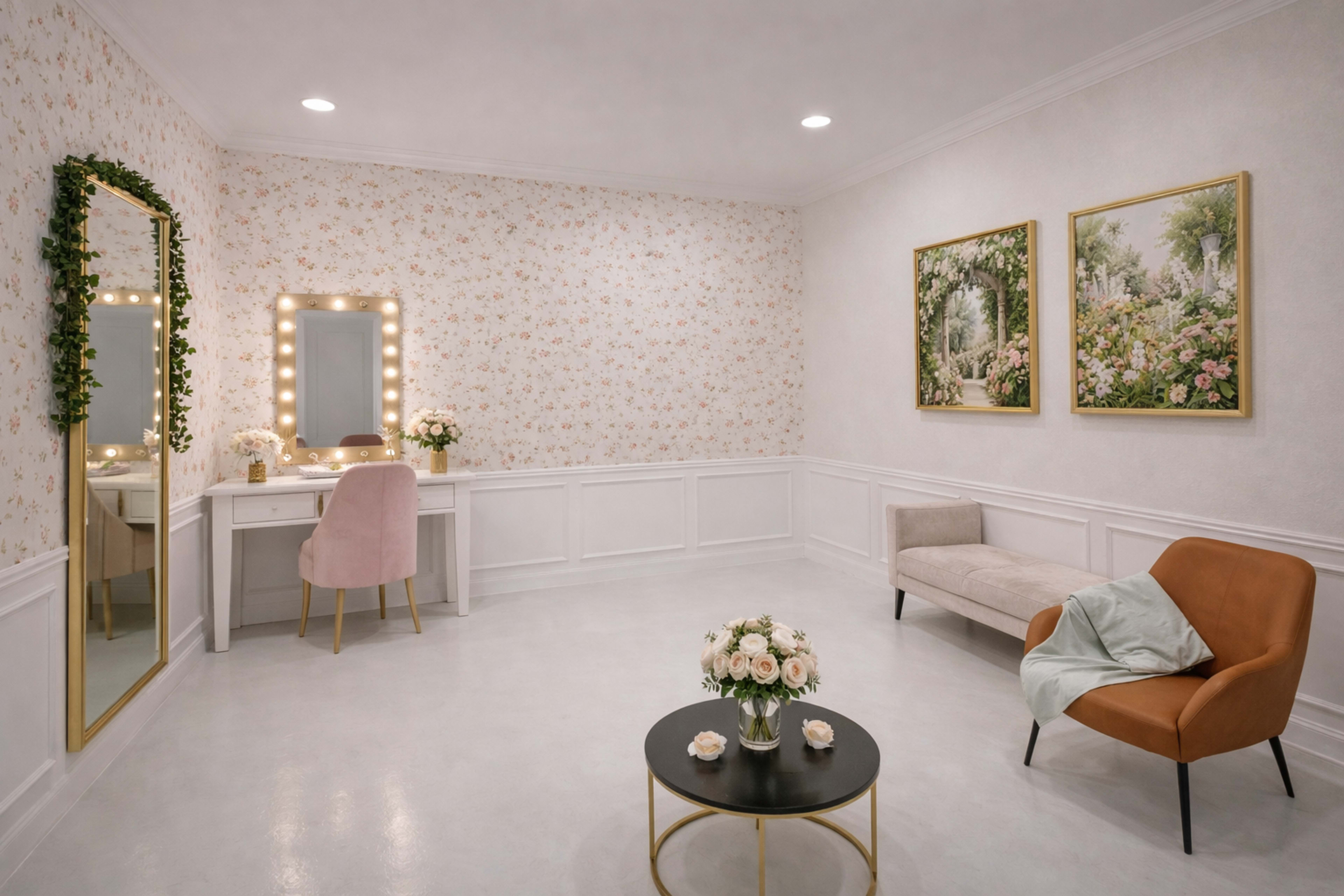 The image shows a well-lit, elegantly decorated room featuring a floral wallpaper, a vanity with a mirror and lights, a small table with flowers, and two stylish chairs facing each other.