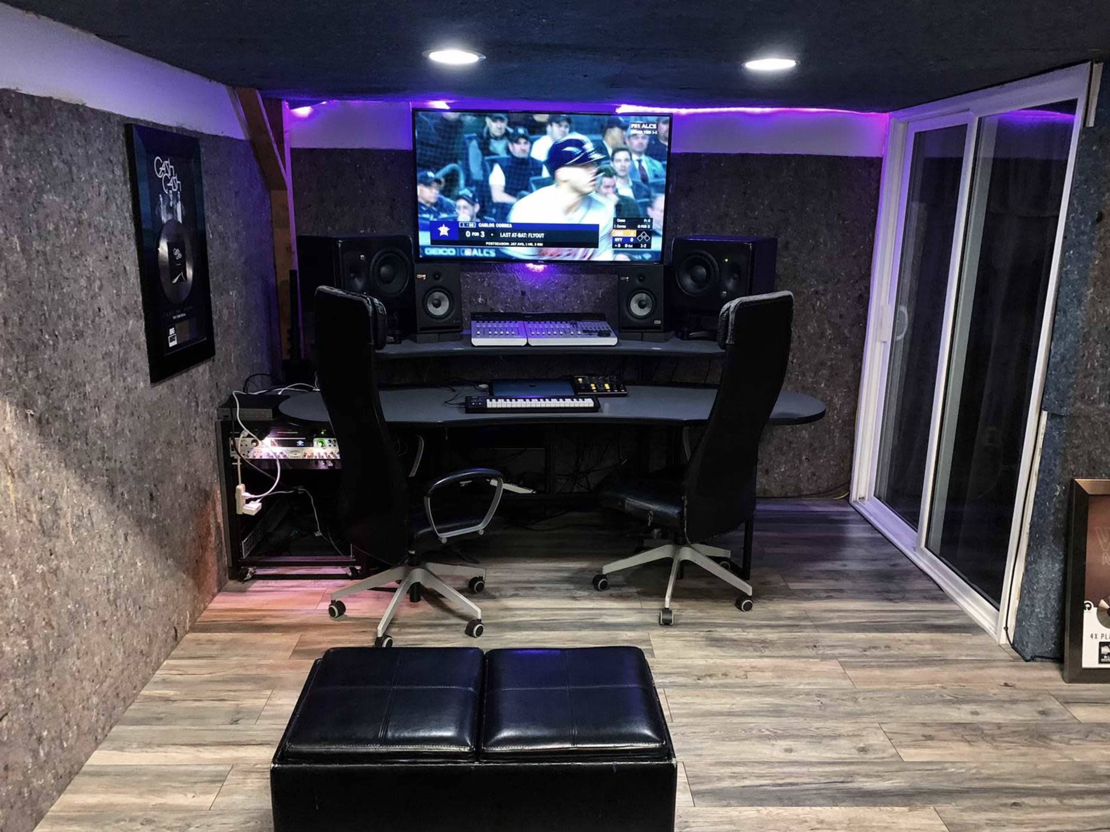 The image shows a modern music production studio featuring a central desk with audio equipment, two large speakers, and a television displaying a sports game on the wall.