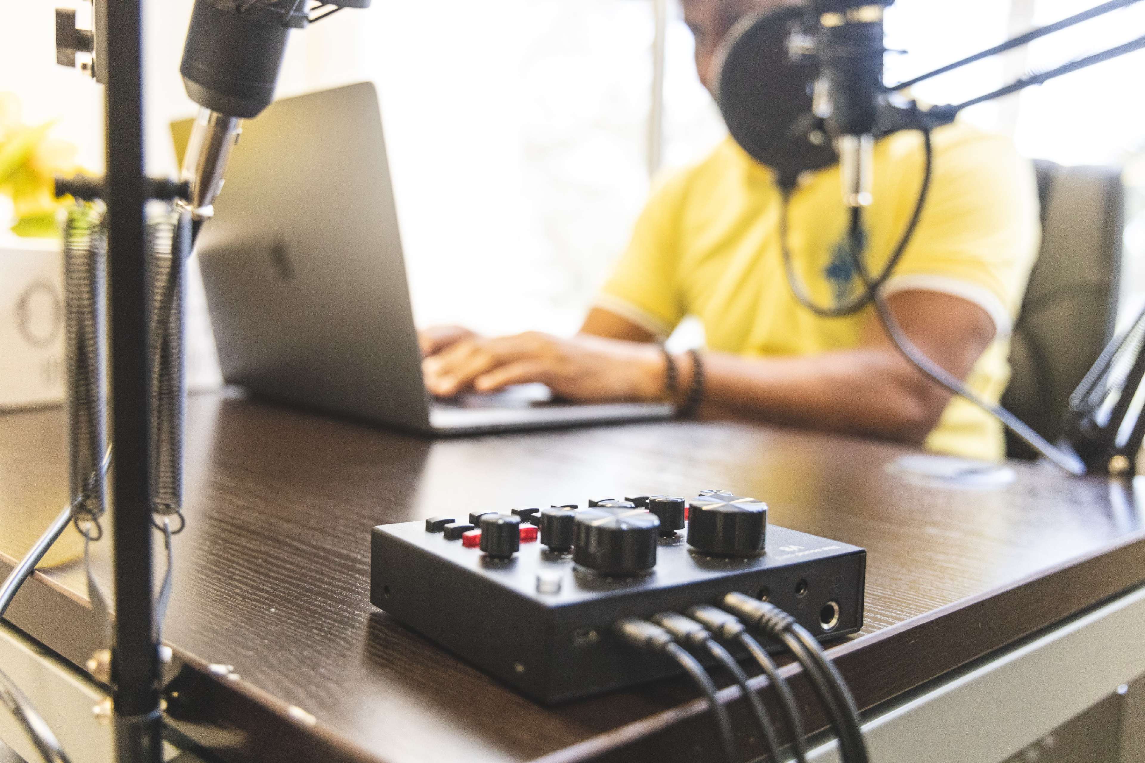 A person sits at a desk using a laptop, with a microphone and audio interface visible in the foreground.