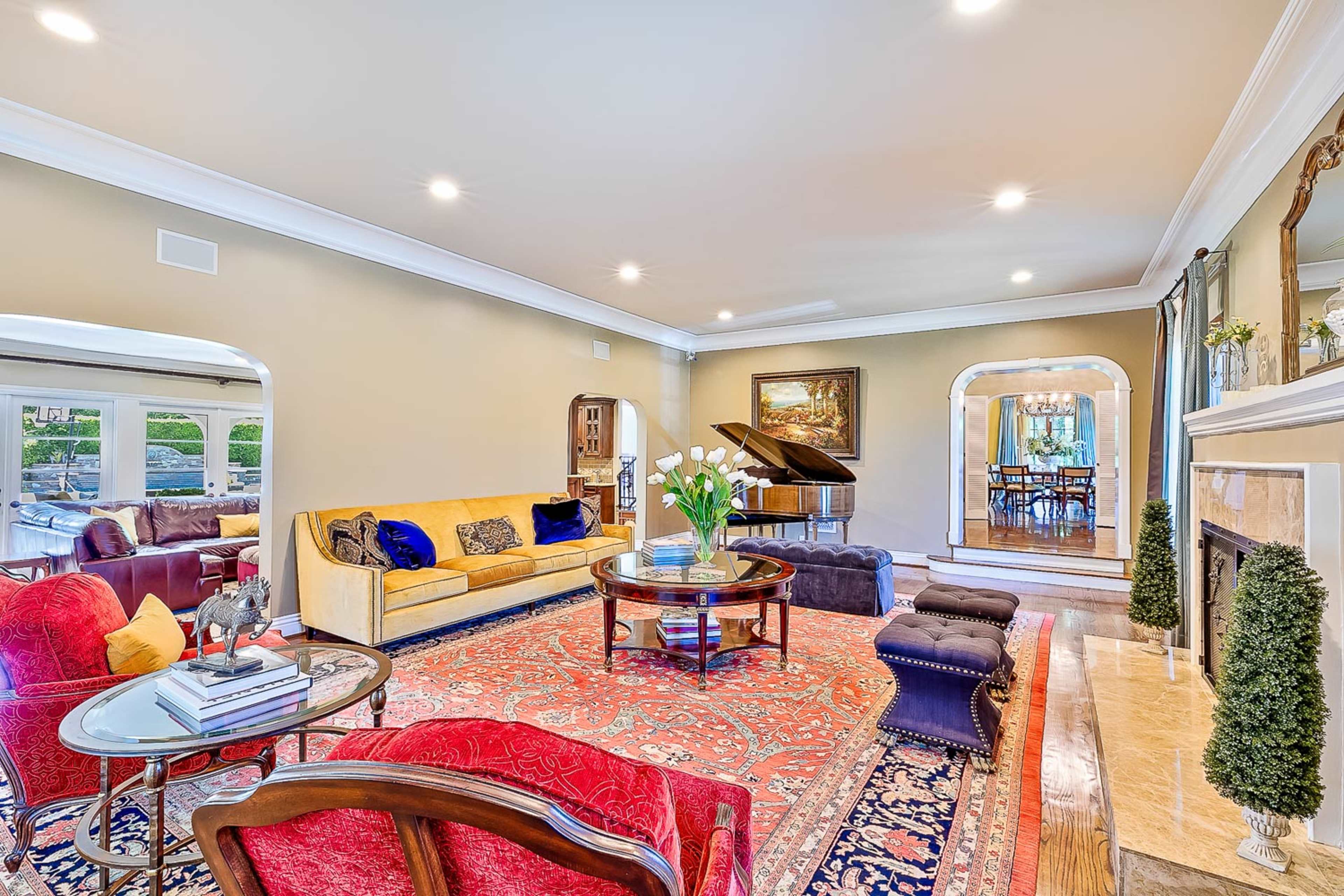 A spacious living room features a grand piano, a yellow sofa, and a decorative rug, surrounded by windows and archways.