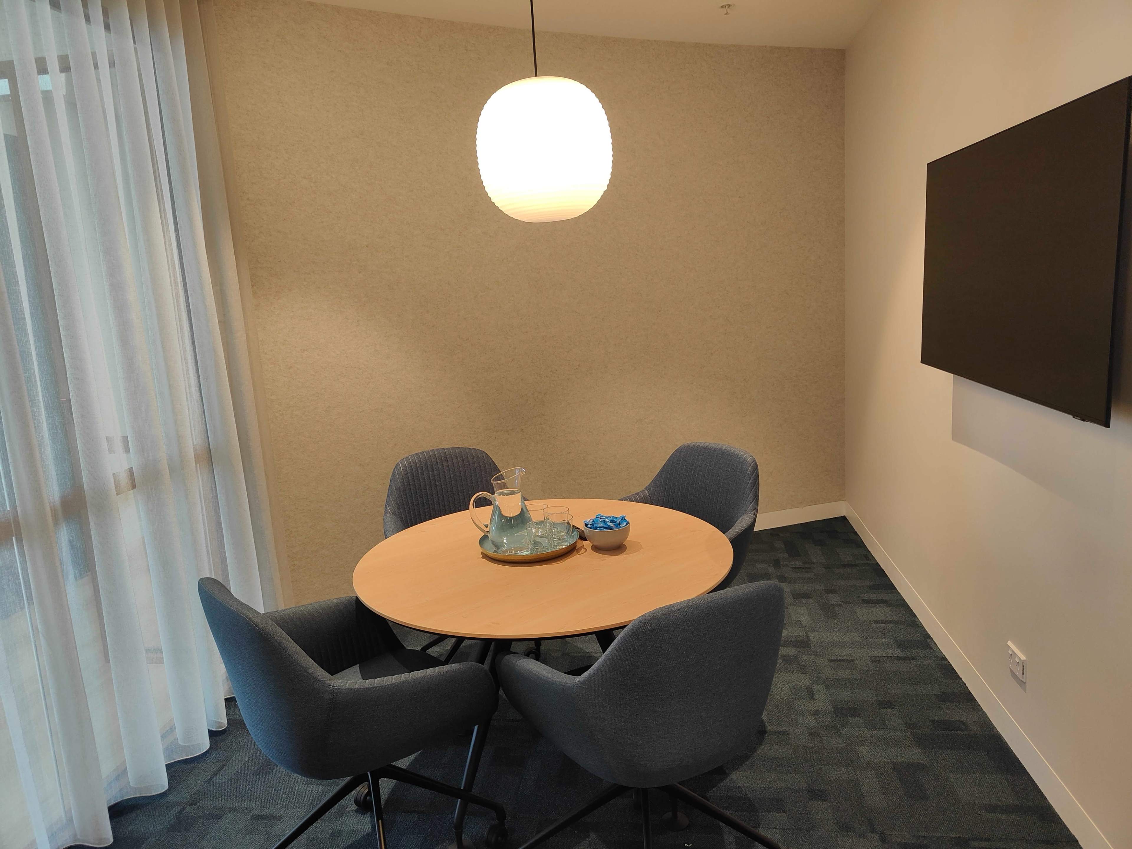 A round wooden table with four gray chairs is set in a corner of a well-lit room, featuring a pitcher and a bowl of snacks, with a large wall-mounted screen on one side.