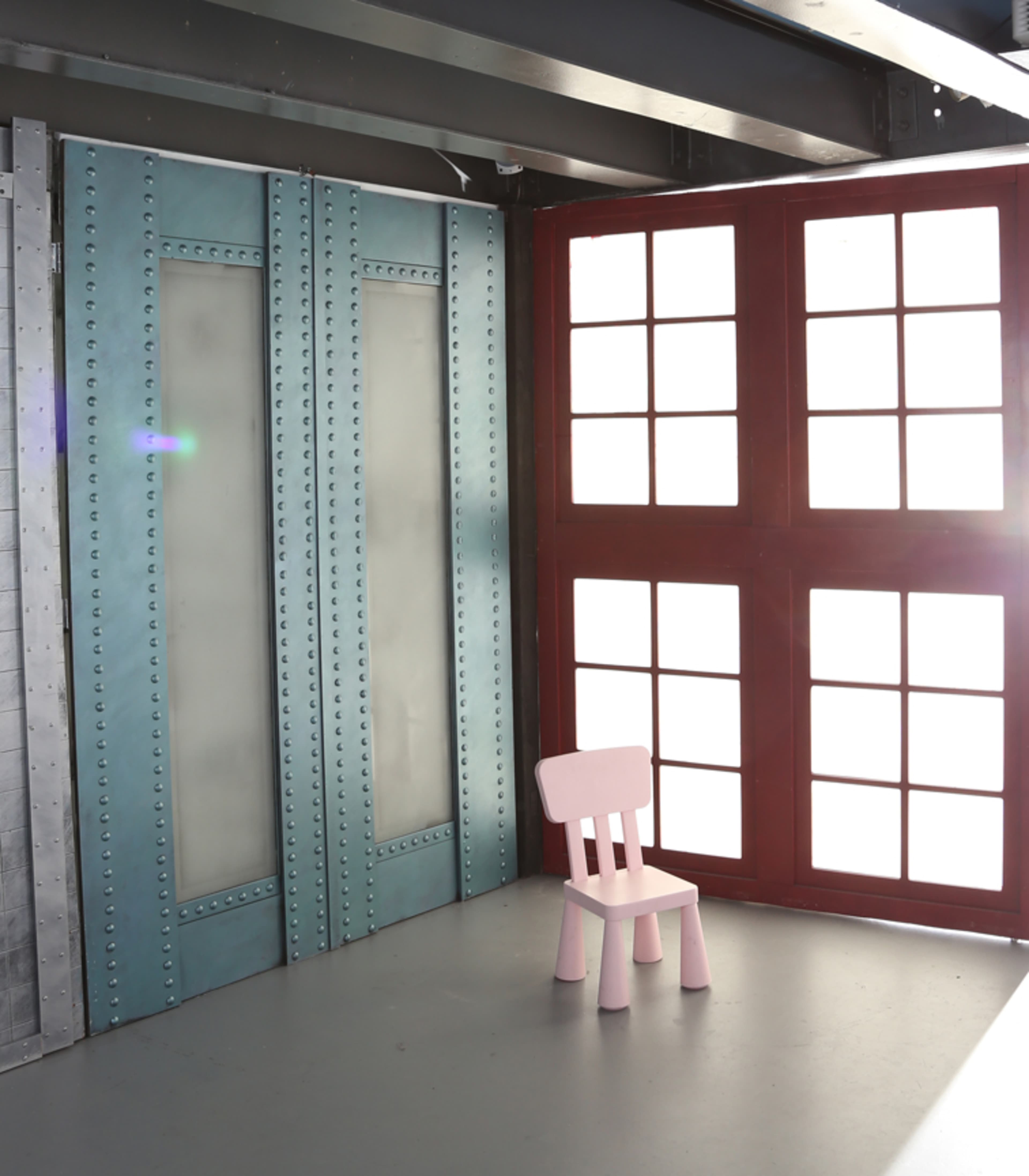 A pink wooden chair stands alone in a well-lit room with large windows and metallic blue walls.
