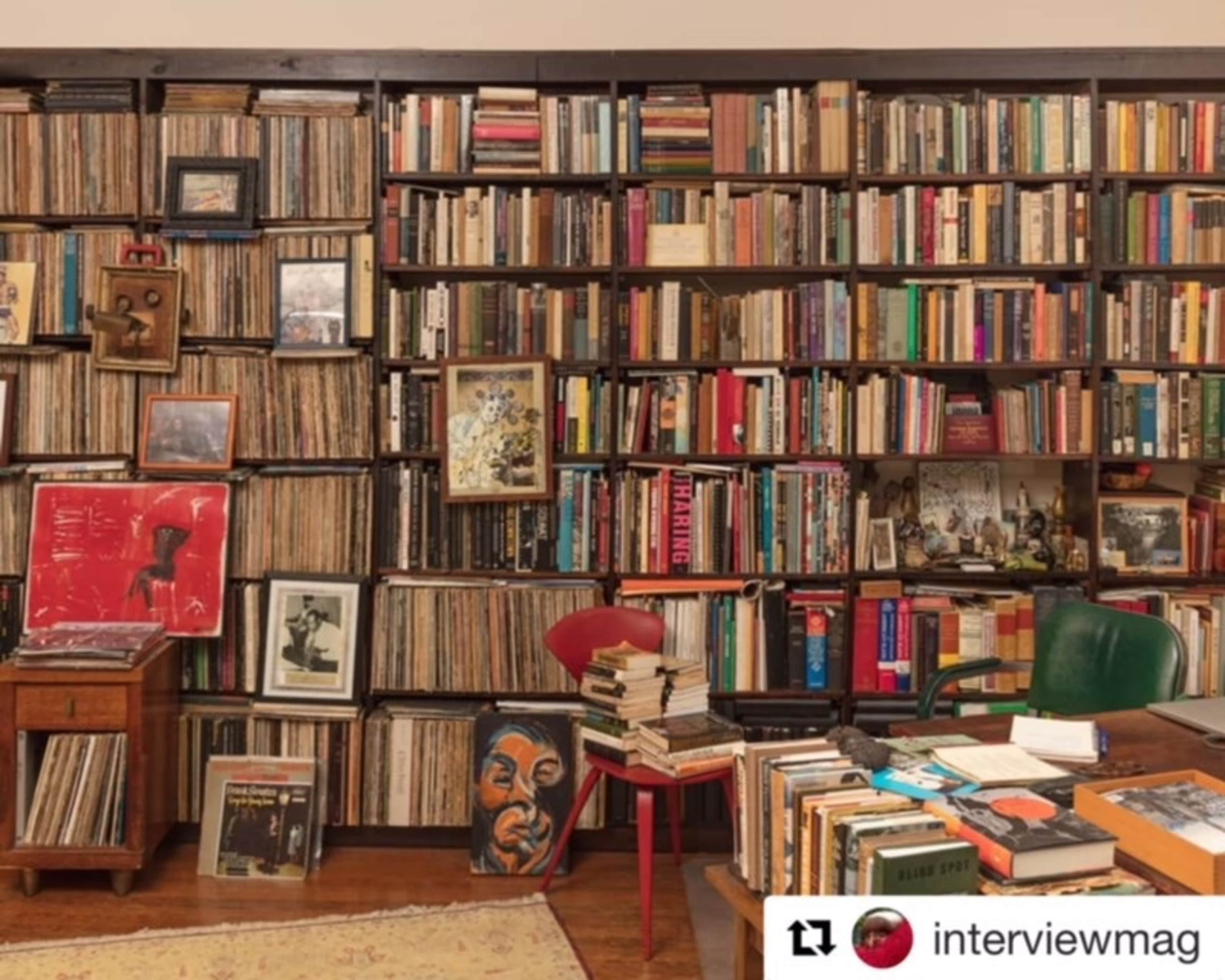 The image shows a wooden bookshelf filled with various books and vinyl records, flanked by a small table stacked with more books and two chairs.