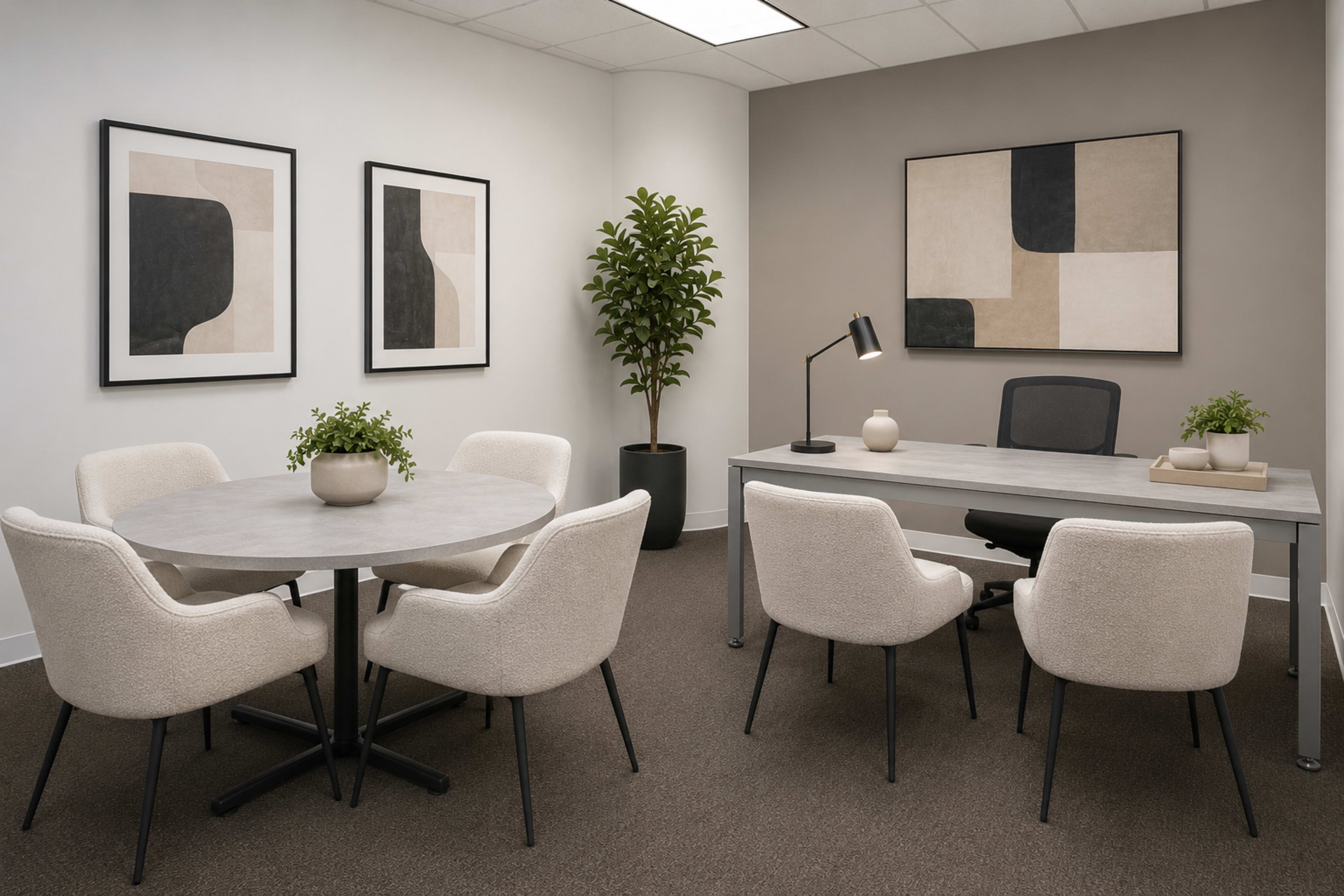 A modern office space features a round dining table with four chairs, a desk with a chair, and artwork on the walls, complemented by a potted plant.