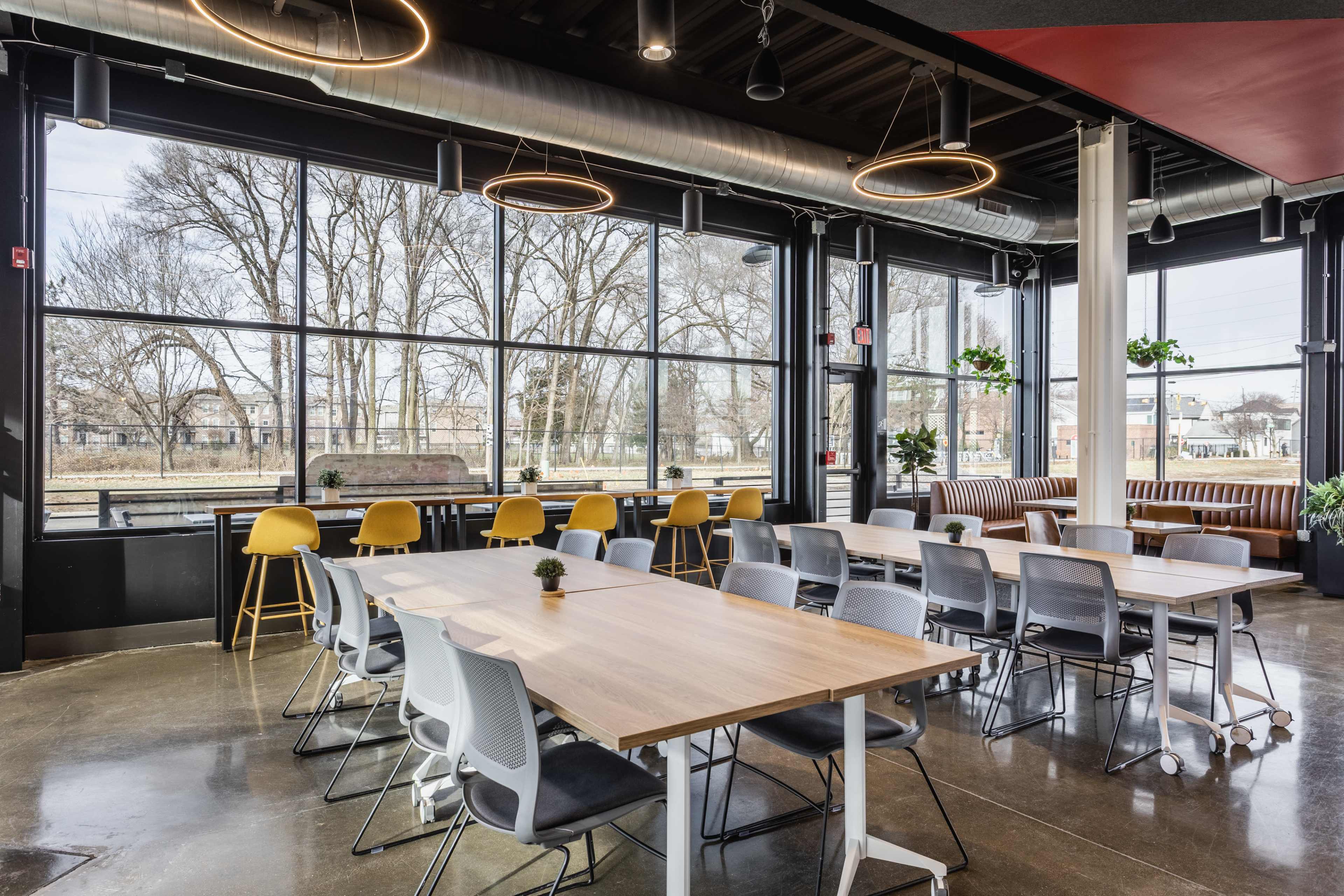 The image shows a modern dining space with large windows, multiple tables, and a few chairs arranged near a seating area.