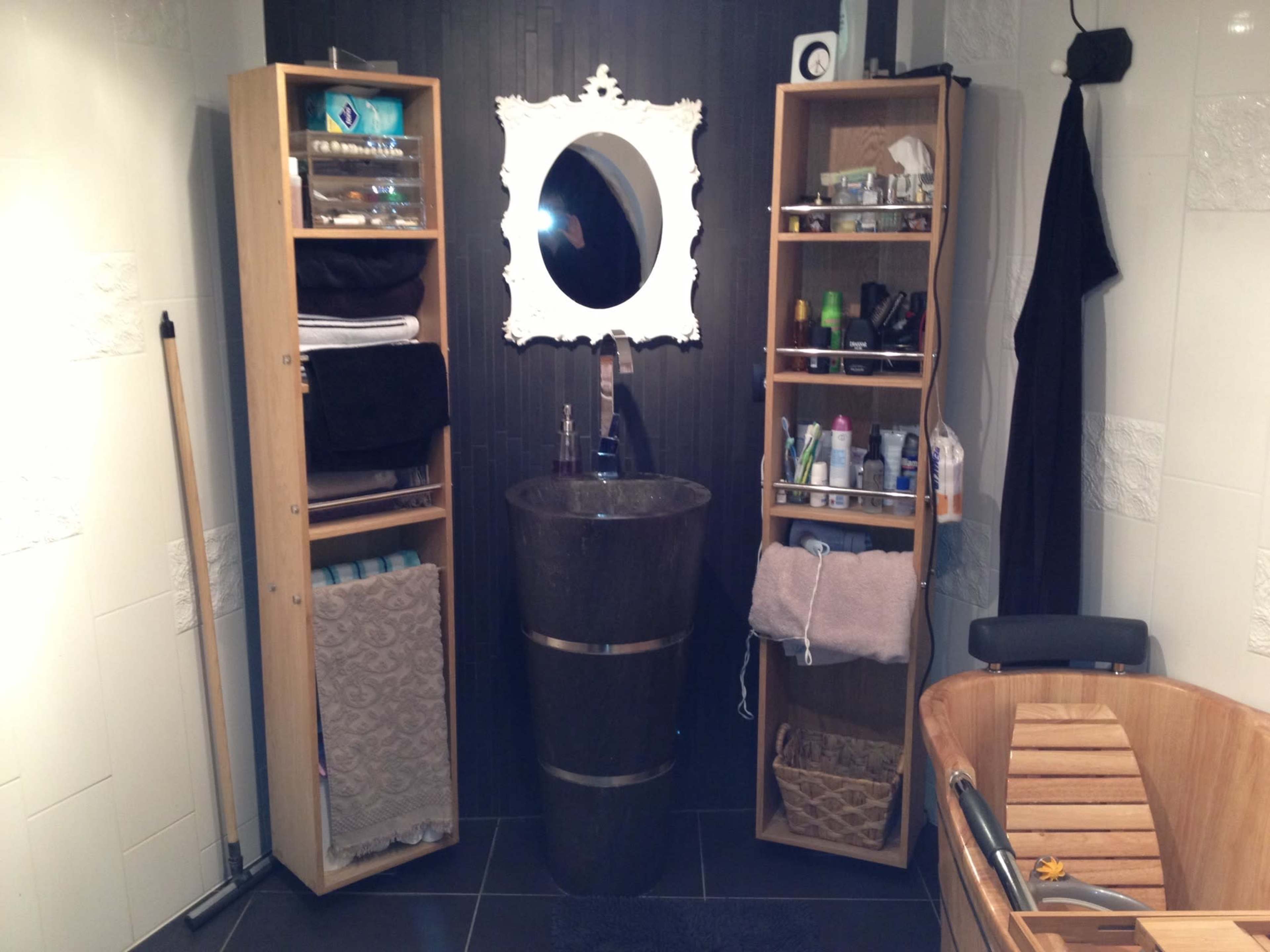 A modern bathroom with a circular stone sink, a wall-mounted mirror in an ornate frame, and wooden shelving units filled with various toiletries.
