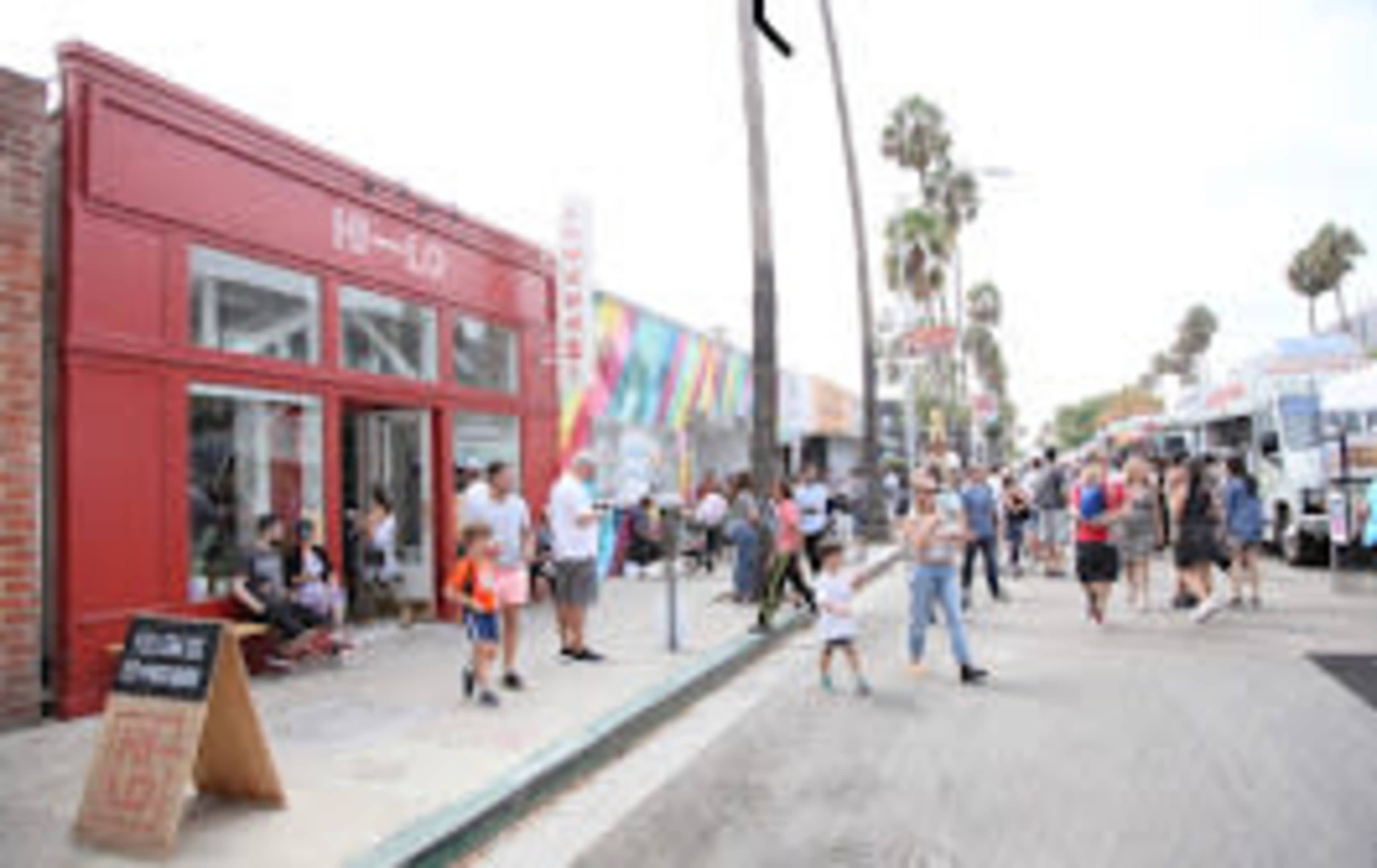 A bustling street scene shows people walking along a road lined with shops and food trucks, with palm trees in the background.