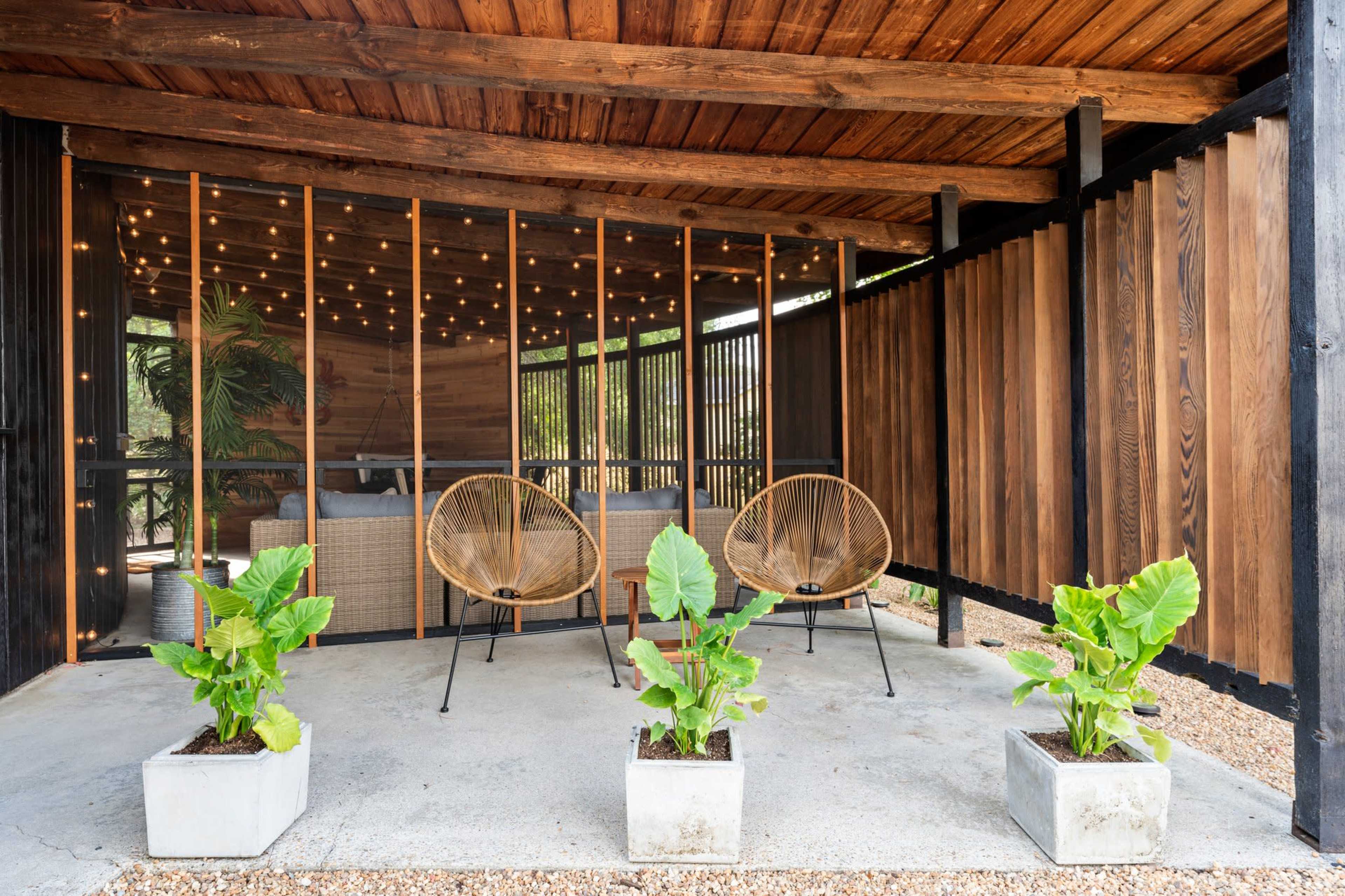 The image shows a covered outdoor seating area with two chairs and three potted plants arranged on a concrete surface.