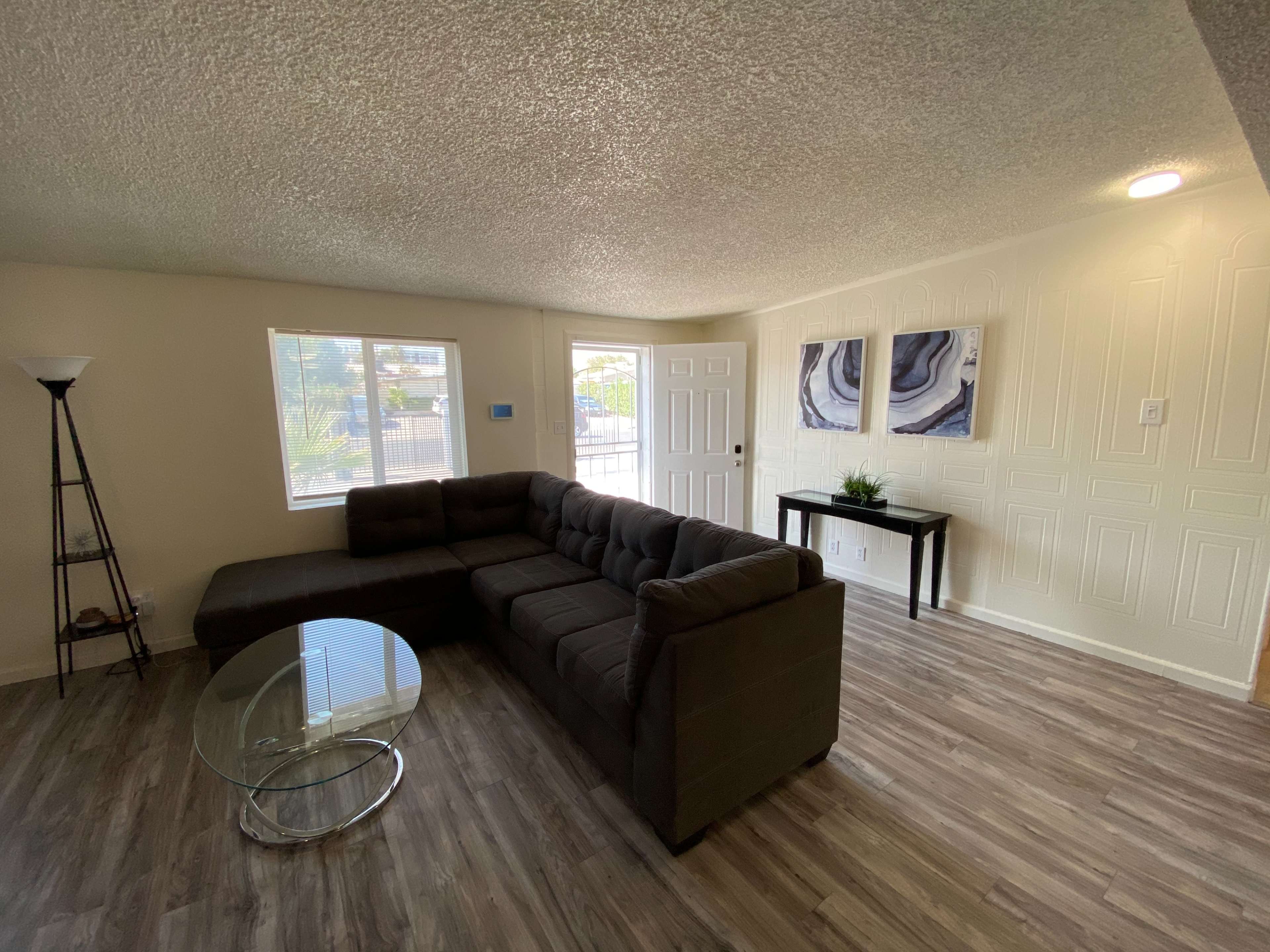 A well-lit living room featuring a large sectional sofa, a glass coffee table, and a console table, with windows providing natural light and a view outside.
