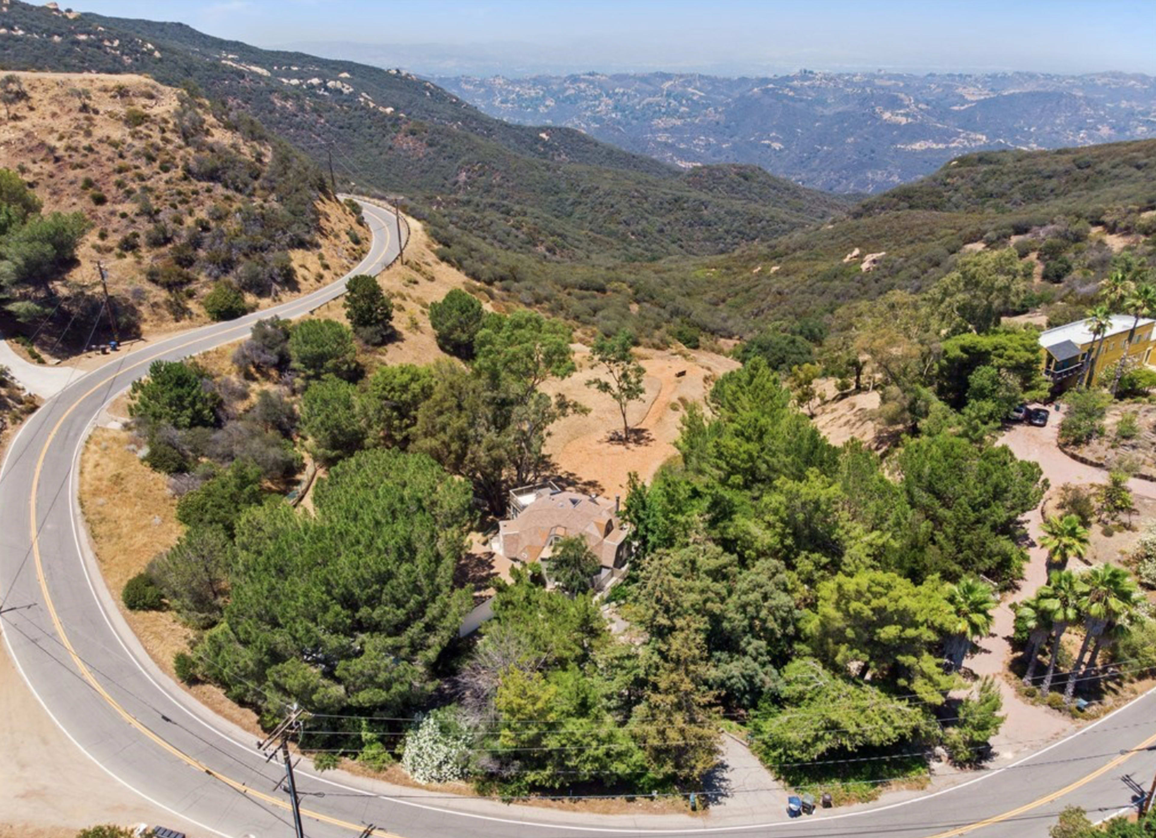 The image shows a winding road curving through a mountainous landscape with sparse trees and a few buildings nestled among the hills.