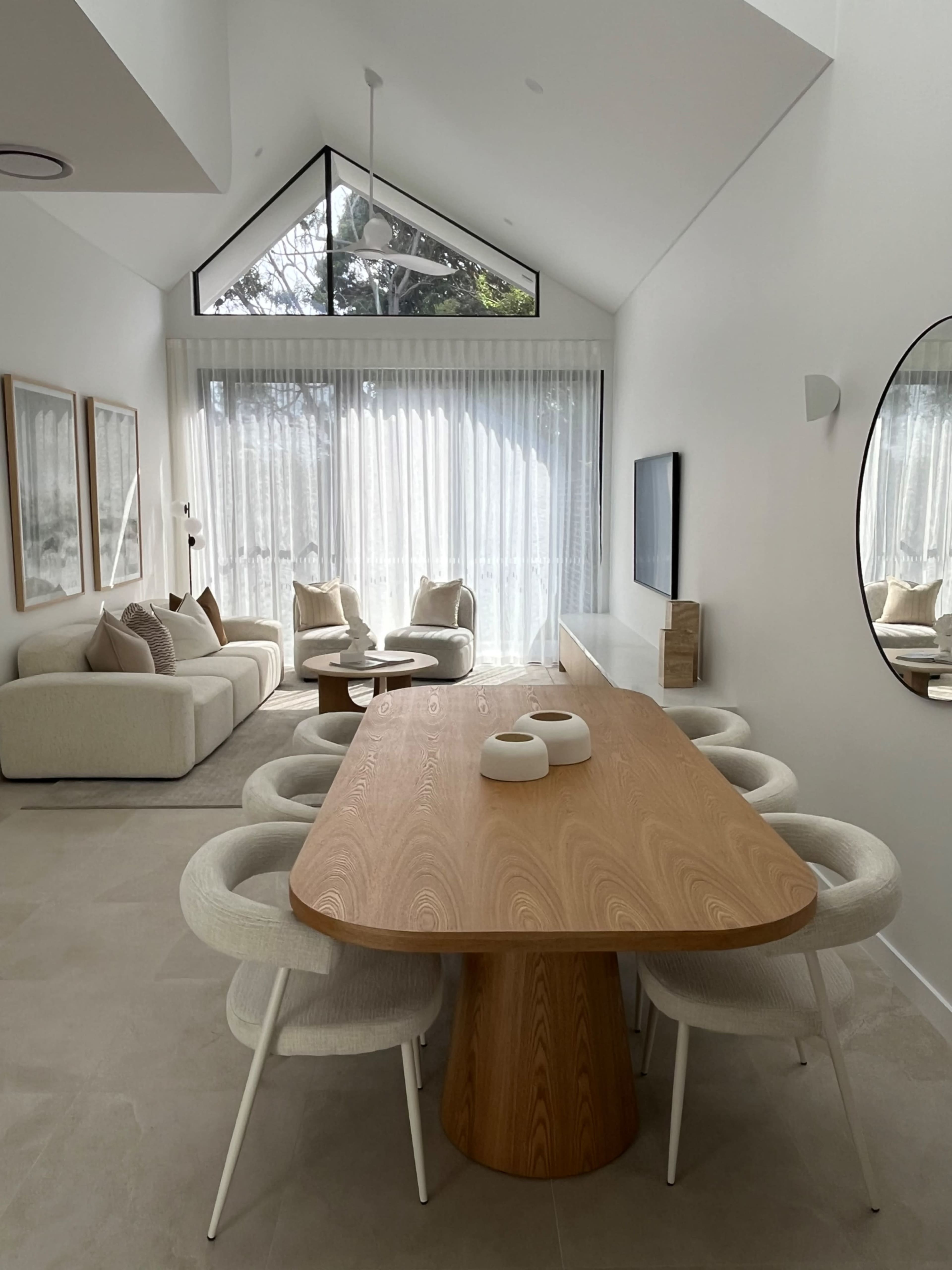 The image depicts a modern dining area featuring a rectangular wooden table surrounded by upholstered chairs, with large windows allowing natural light to enter the space.