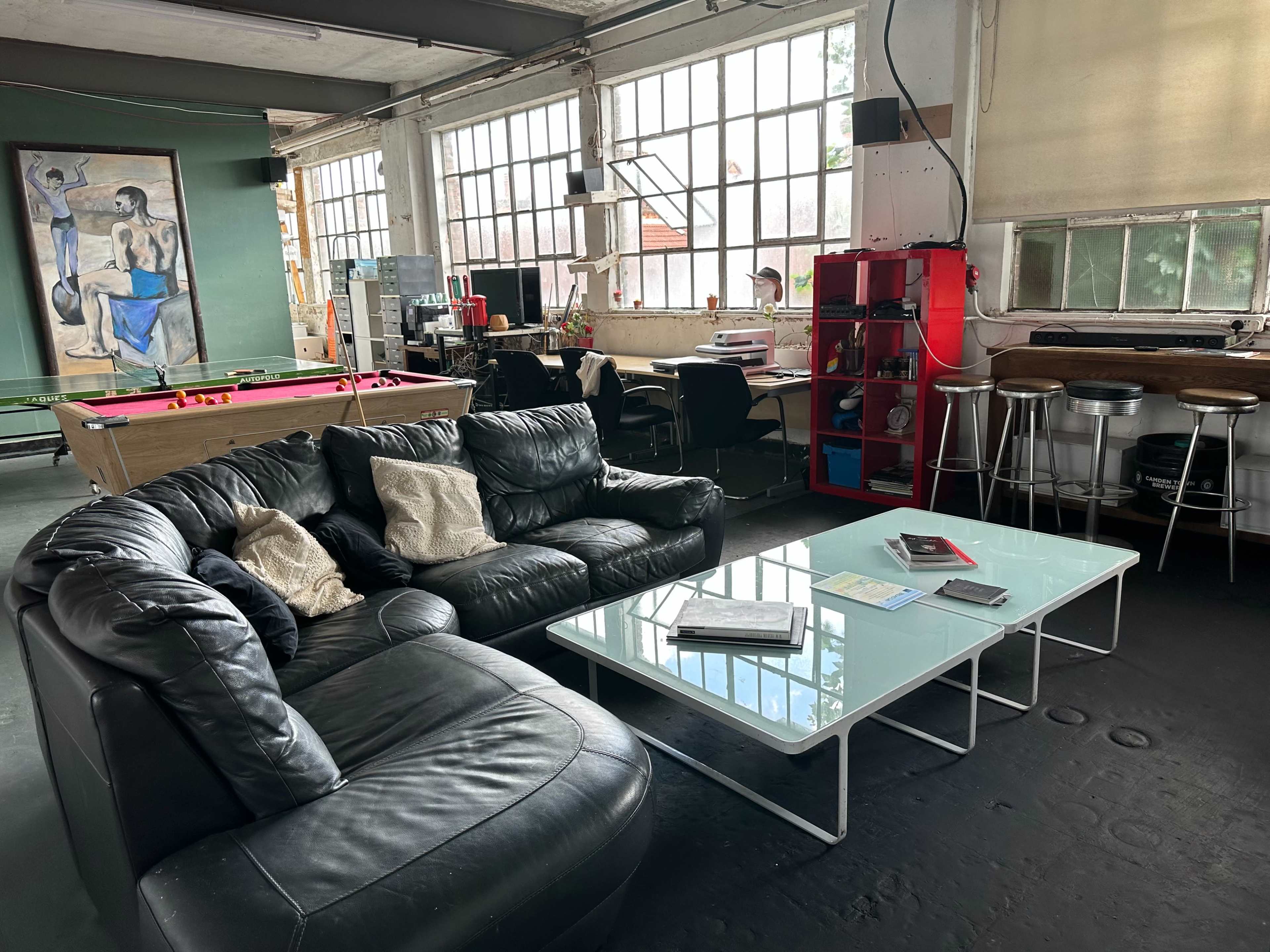 A spacious industrial-inspired room with large windows, featuring a black leather sofa, a glass coffee table, and workstations in the background.