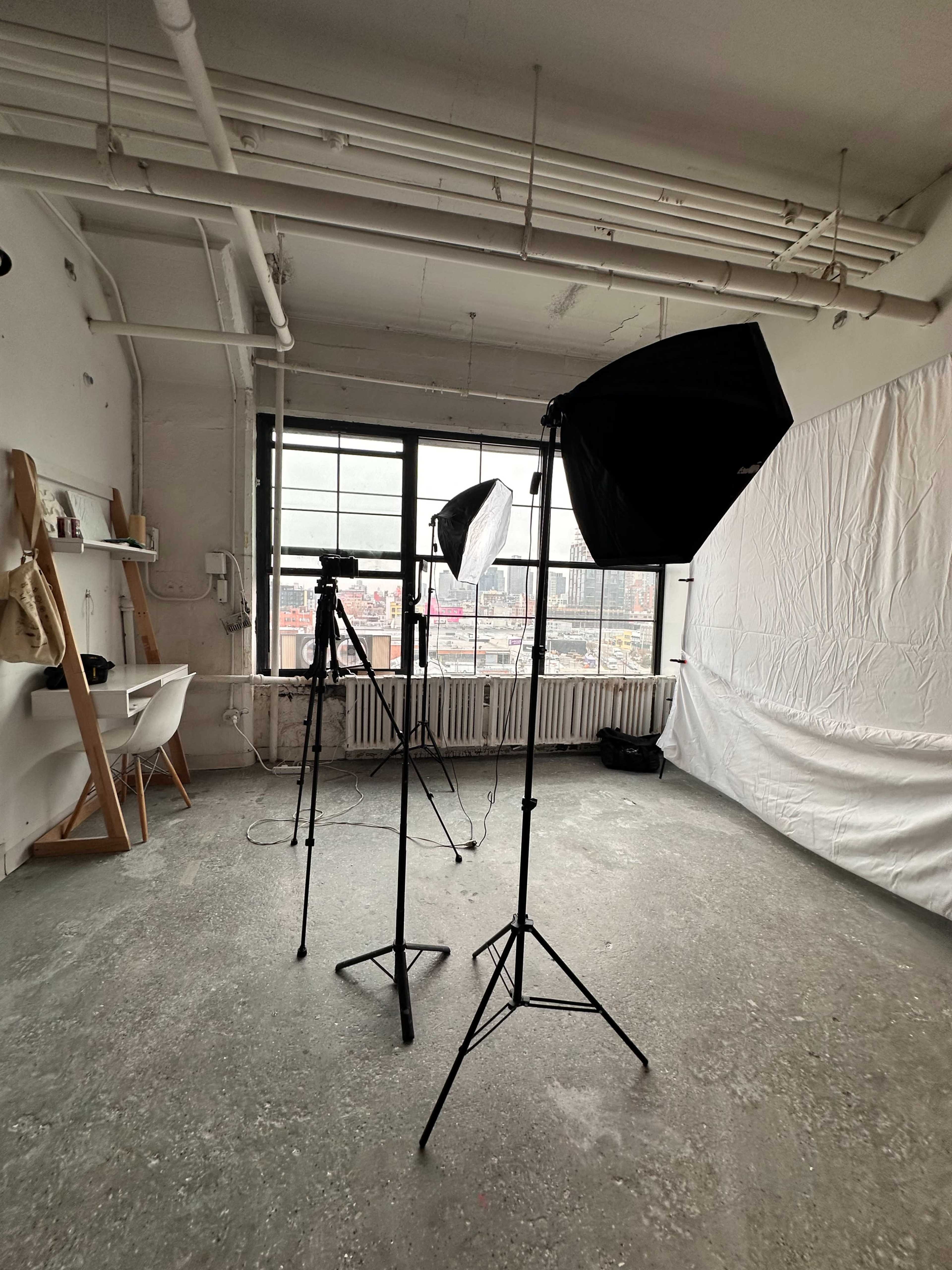 The image shows a studio space with three light stands, a camera mounted on a tripod, and a white backdrop positioned against a large window.