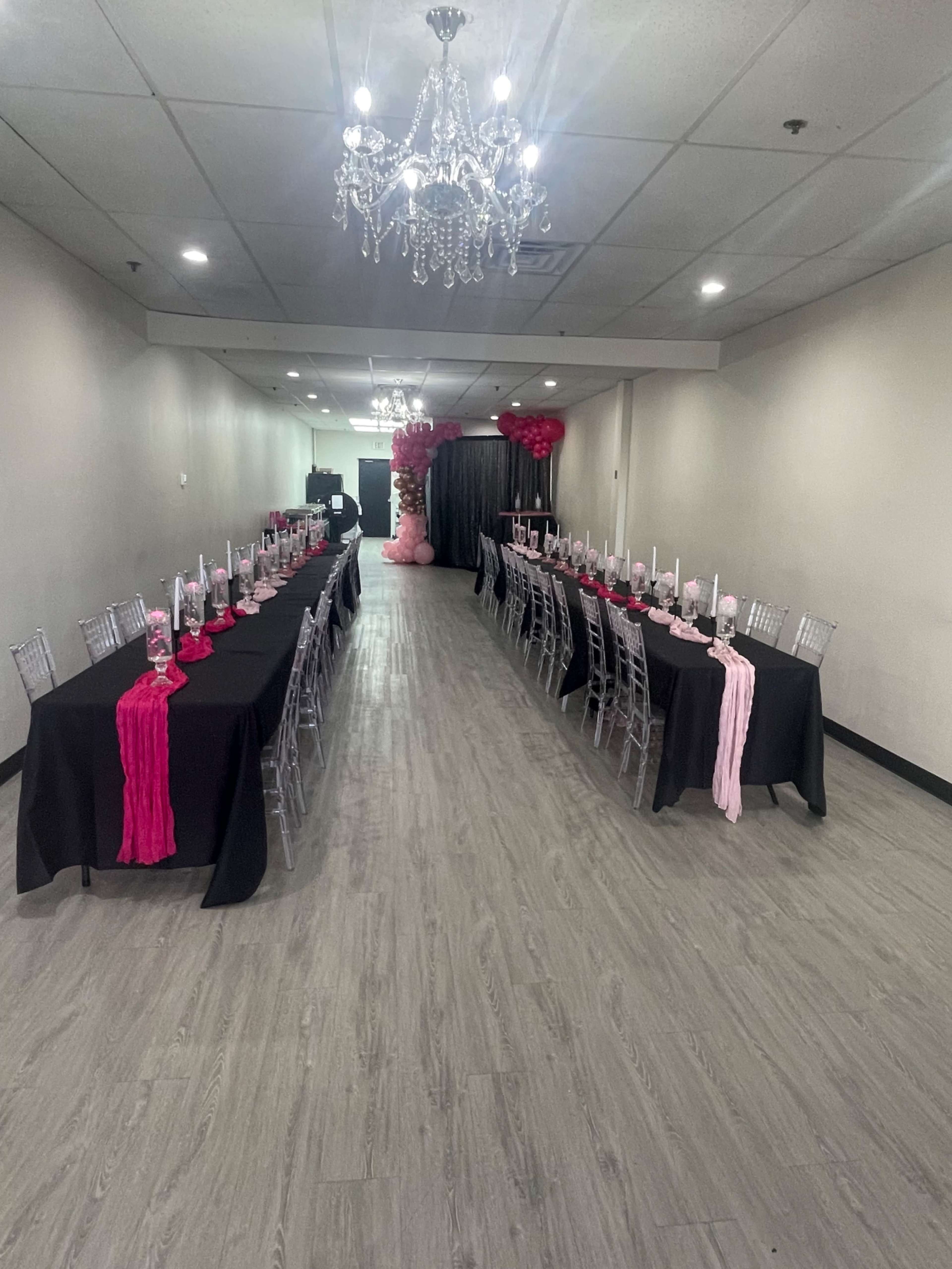 A long banquet hall features two rows of elegantly set tables with black tablecloths and decorative pink accents.