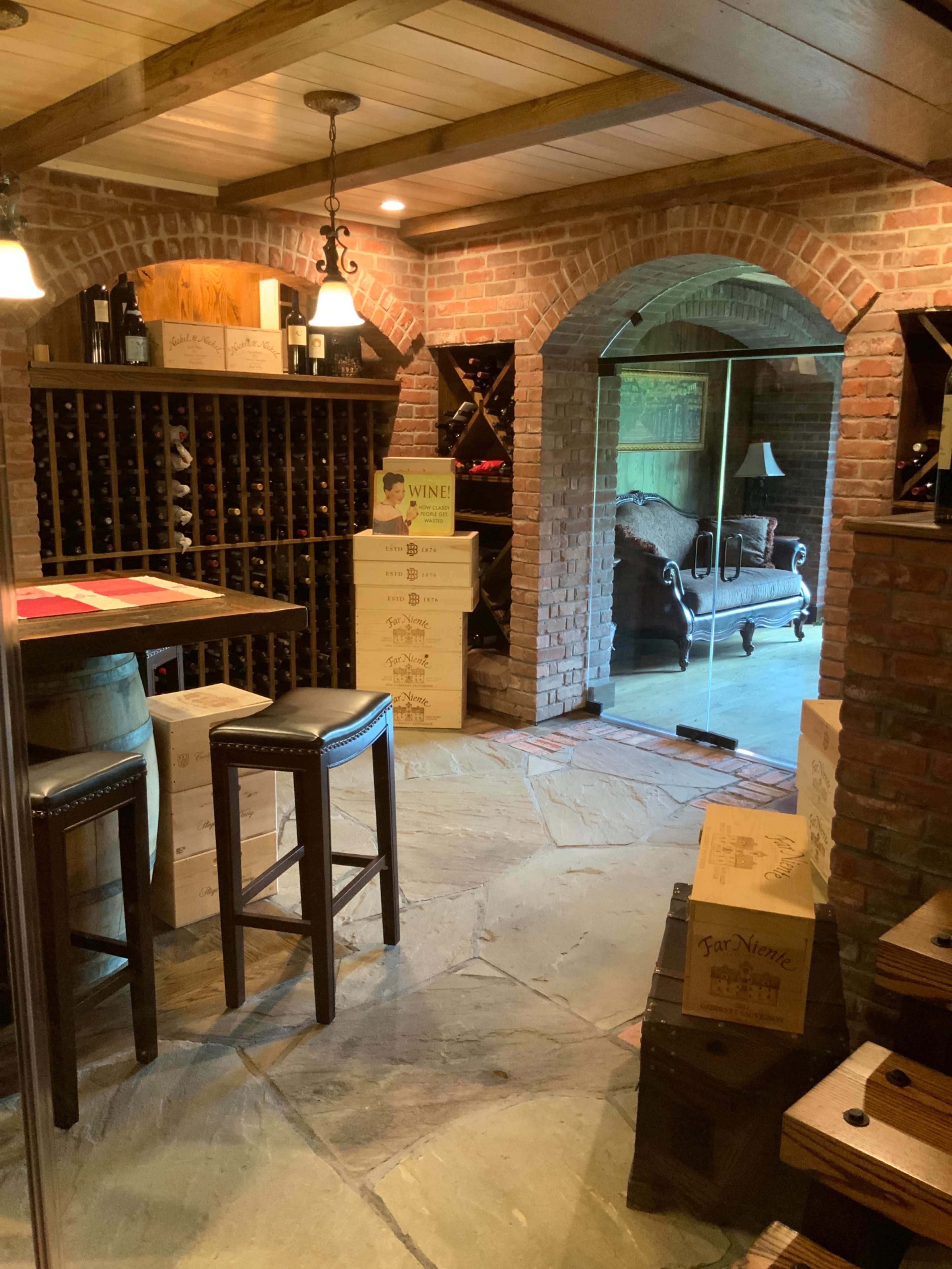 A wine cellar with brick walls, wooden beams, a wine rack, and boxes stacked near a seating area.