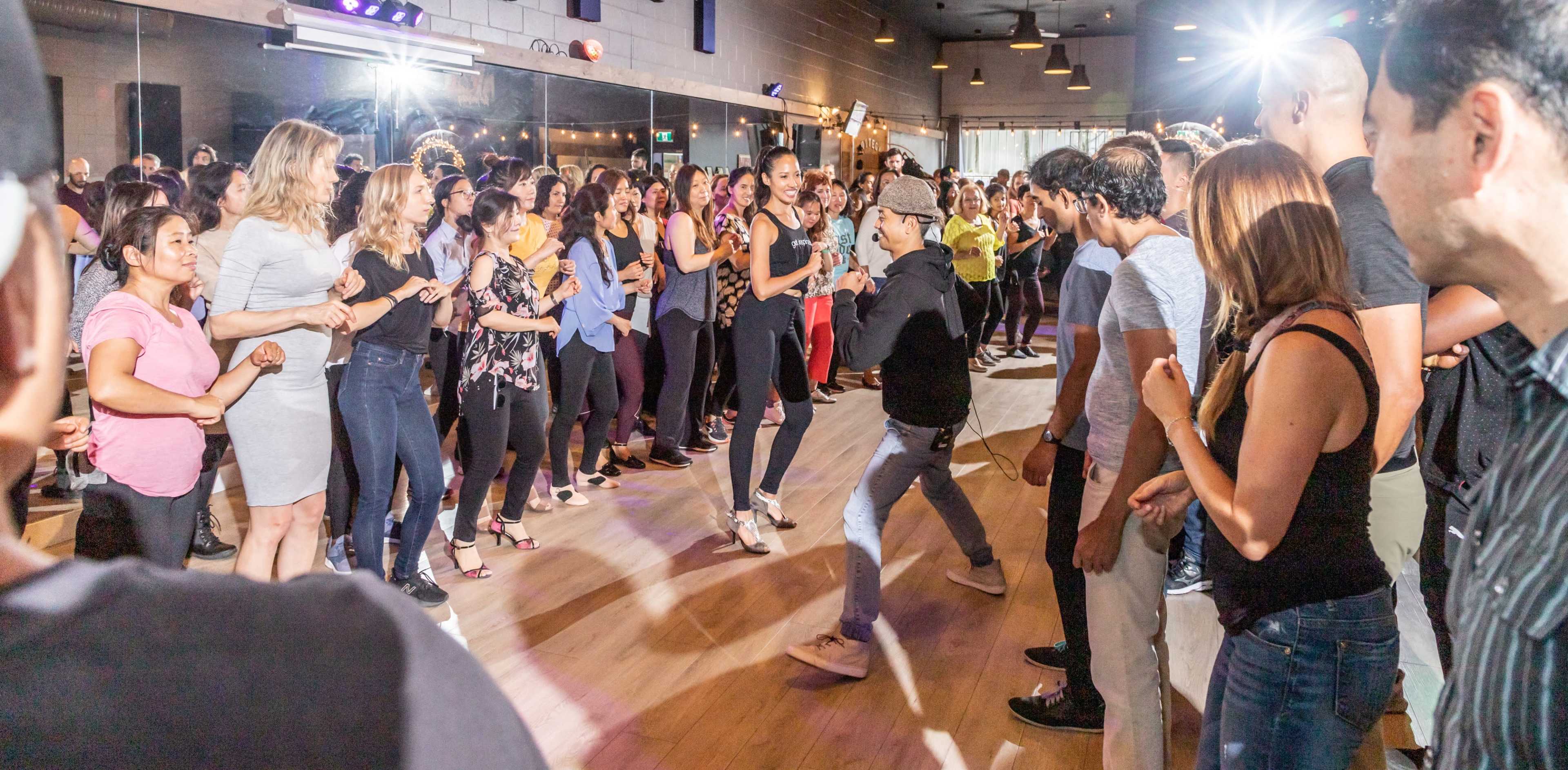 A large group of people dance in a lively studio with lights and a wooden floor.