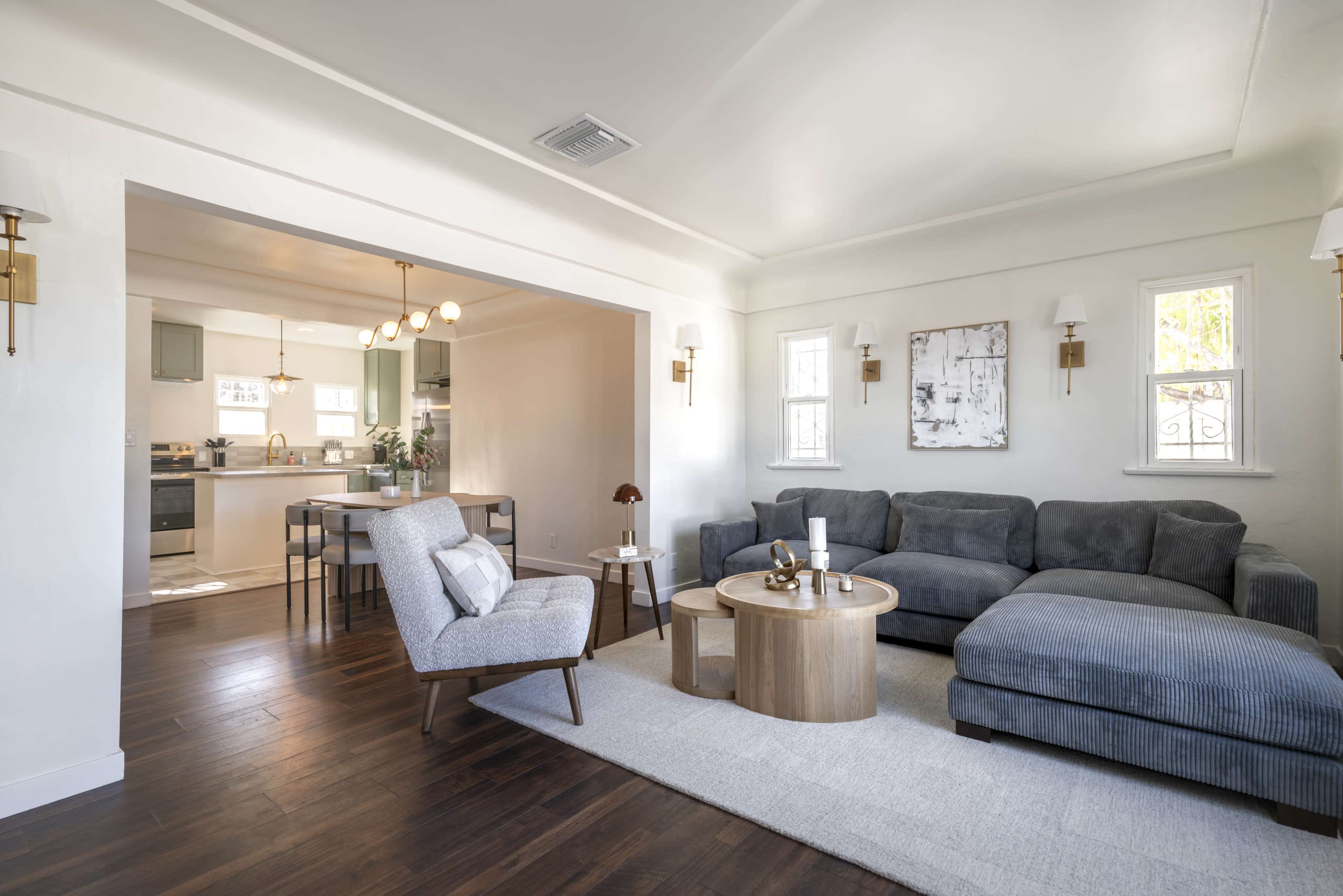 The image shows a modern living room with a gray sectional sofa, a round wooden coffee table, and a cozy armchair, adjacent to a dining area with a table and chairs.