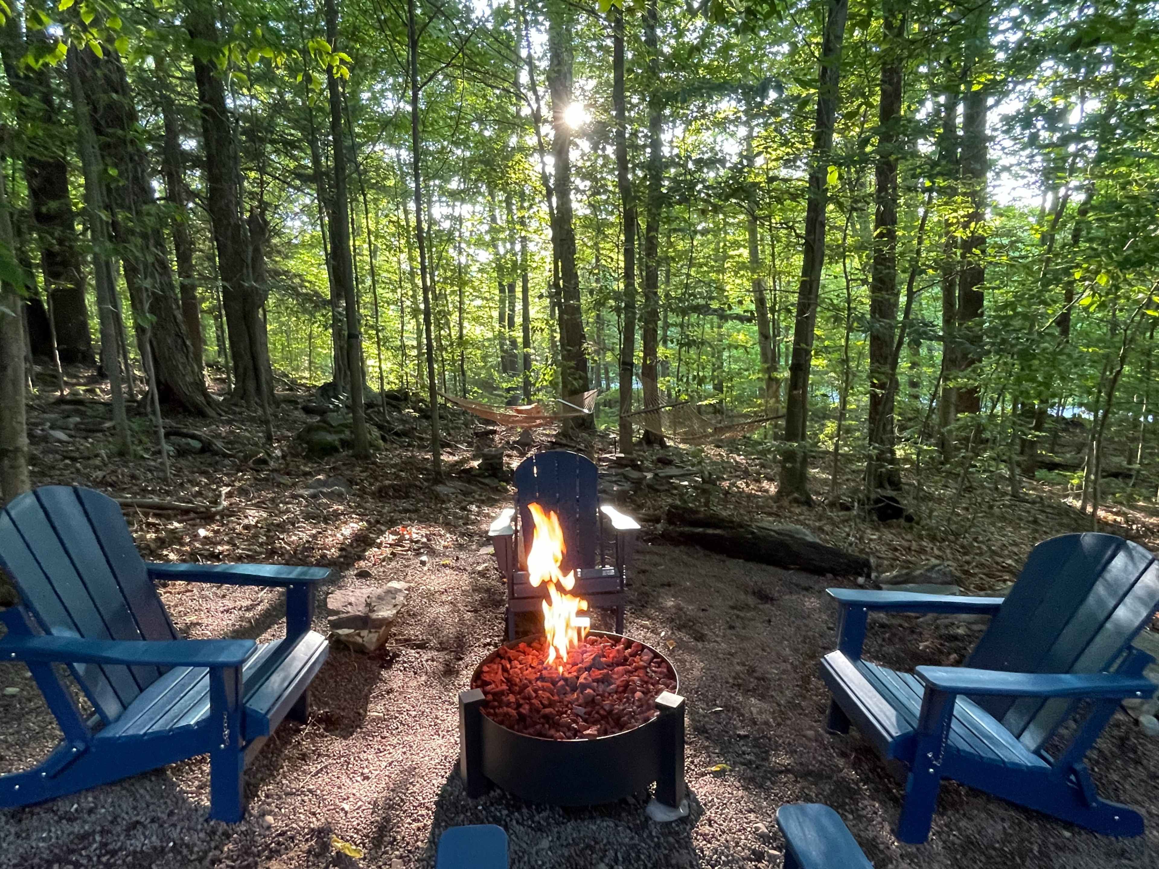 A circular fire pit with a flame surrounded by blue adirondack chairs sits in a forested area with trees and a hammock in the background.