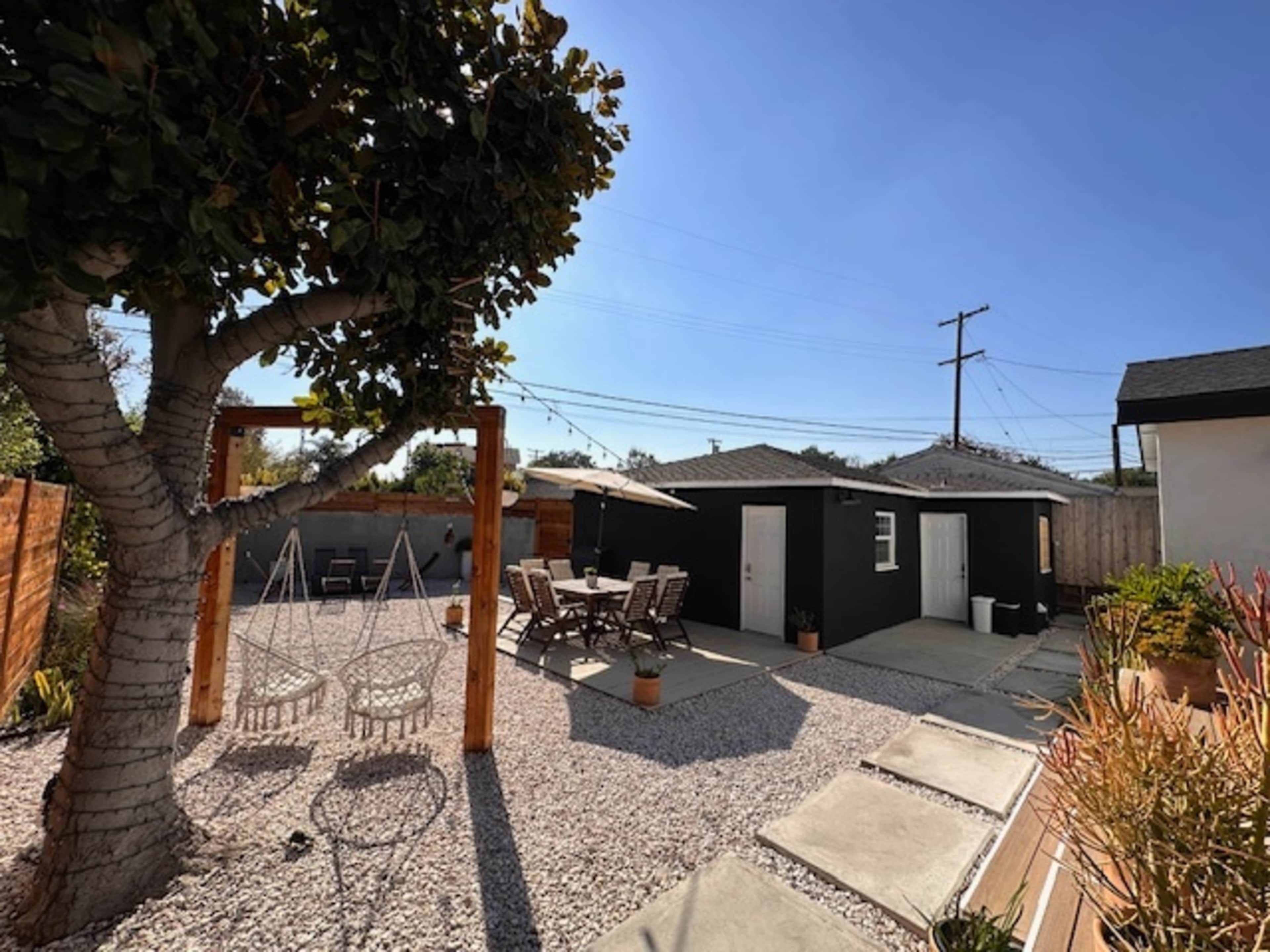 A yard features a tree with swings, a gravel area with outdoor seating, and a black shed in the background under a clear sky.