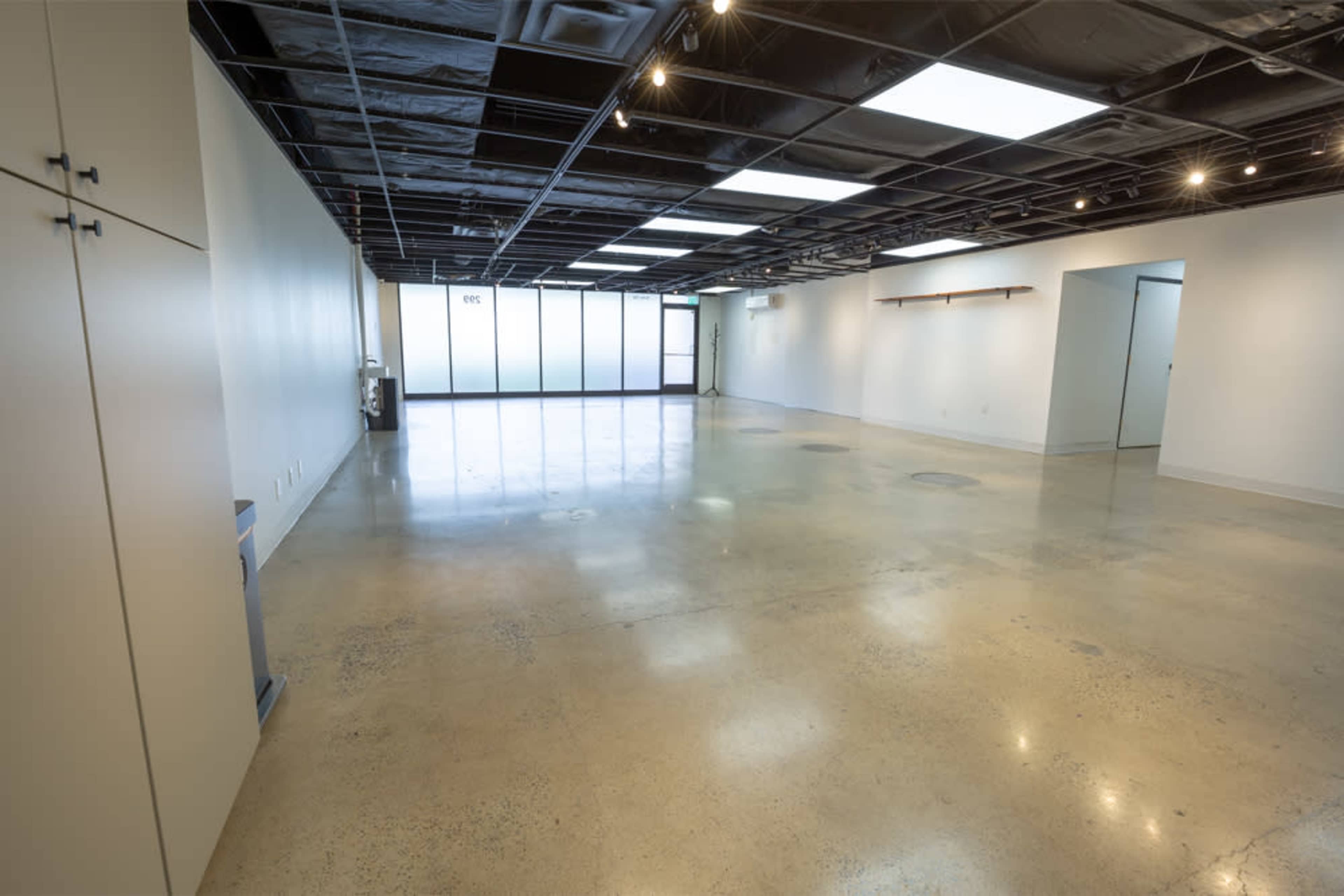 The image shows an empty commercial space with polished concrete floors and white walls, featuring large windows on one side.