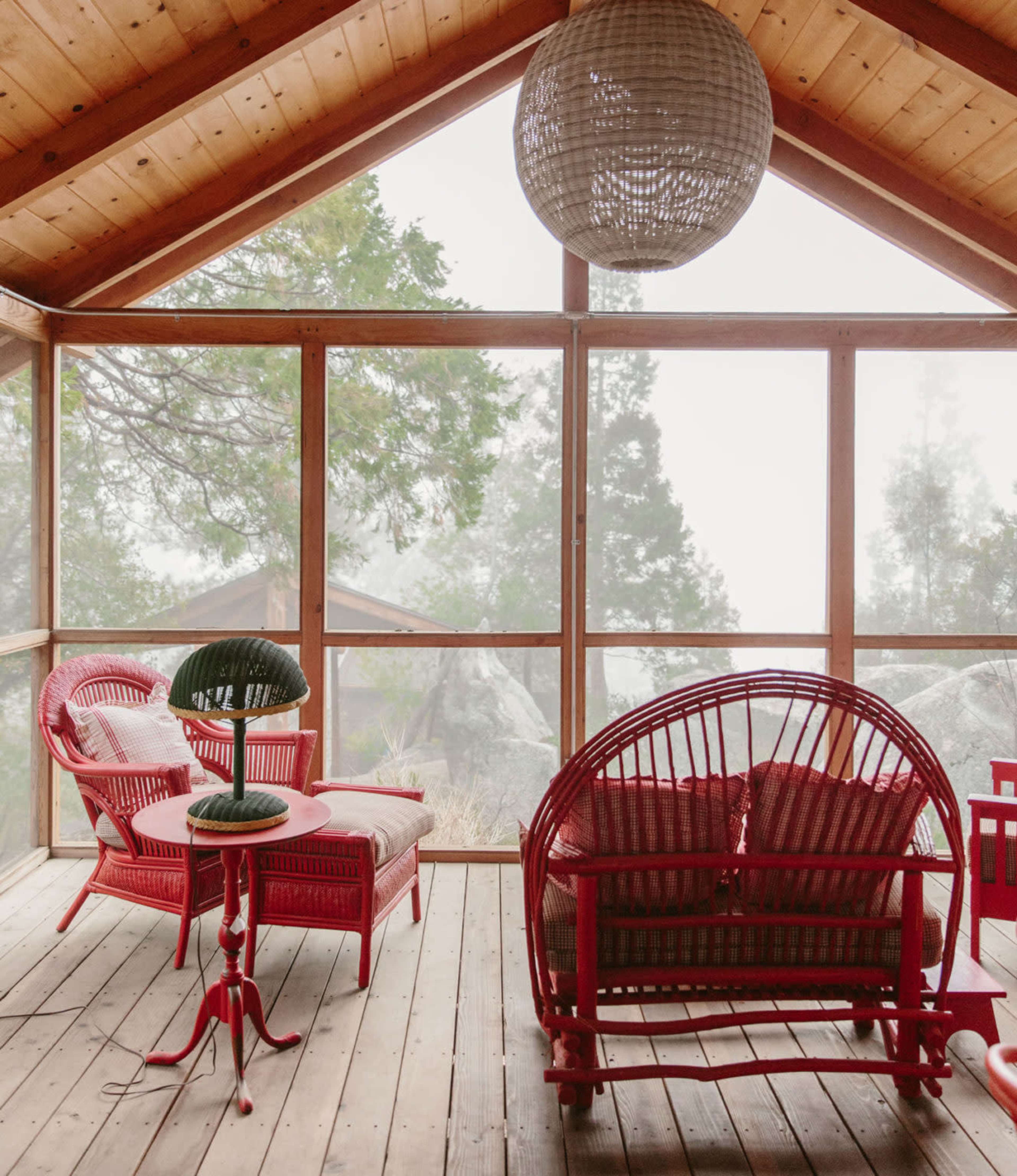 A sunlit screened porch features red wicker furniture and a spherical hanging lamp, with misty trees visible through the windows.