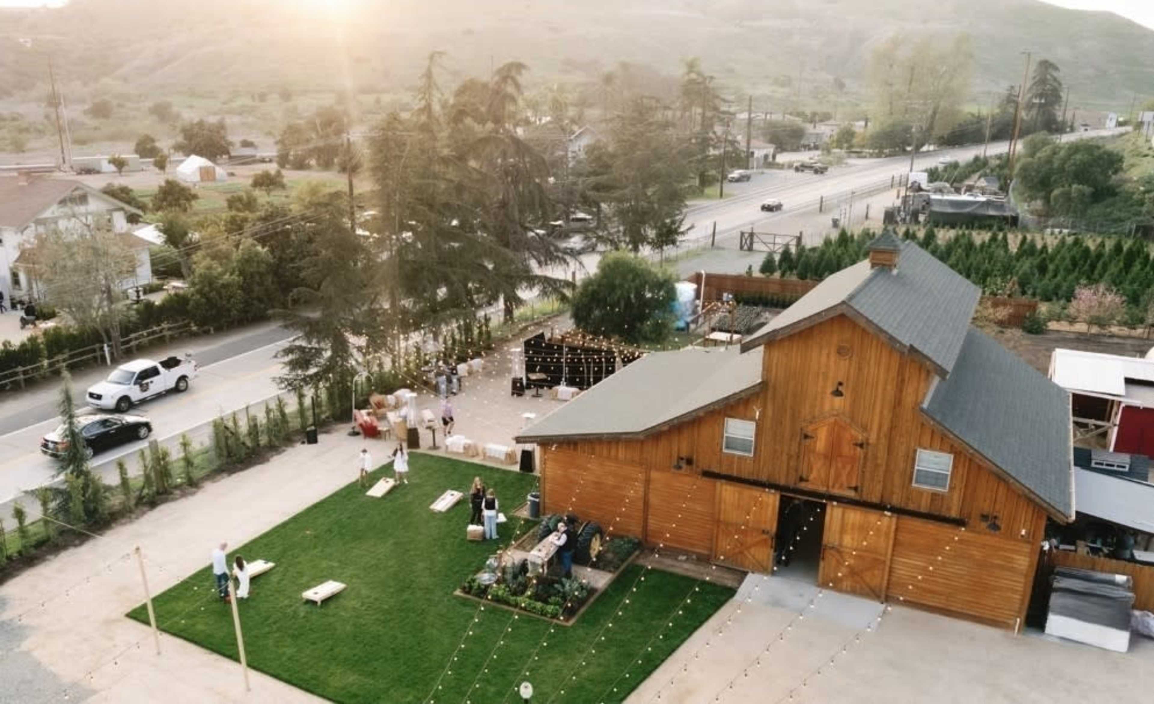 A wooden barn surrounded by green grass and trees is set near a road, with people engaged in various activities outside.