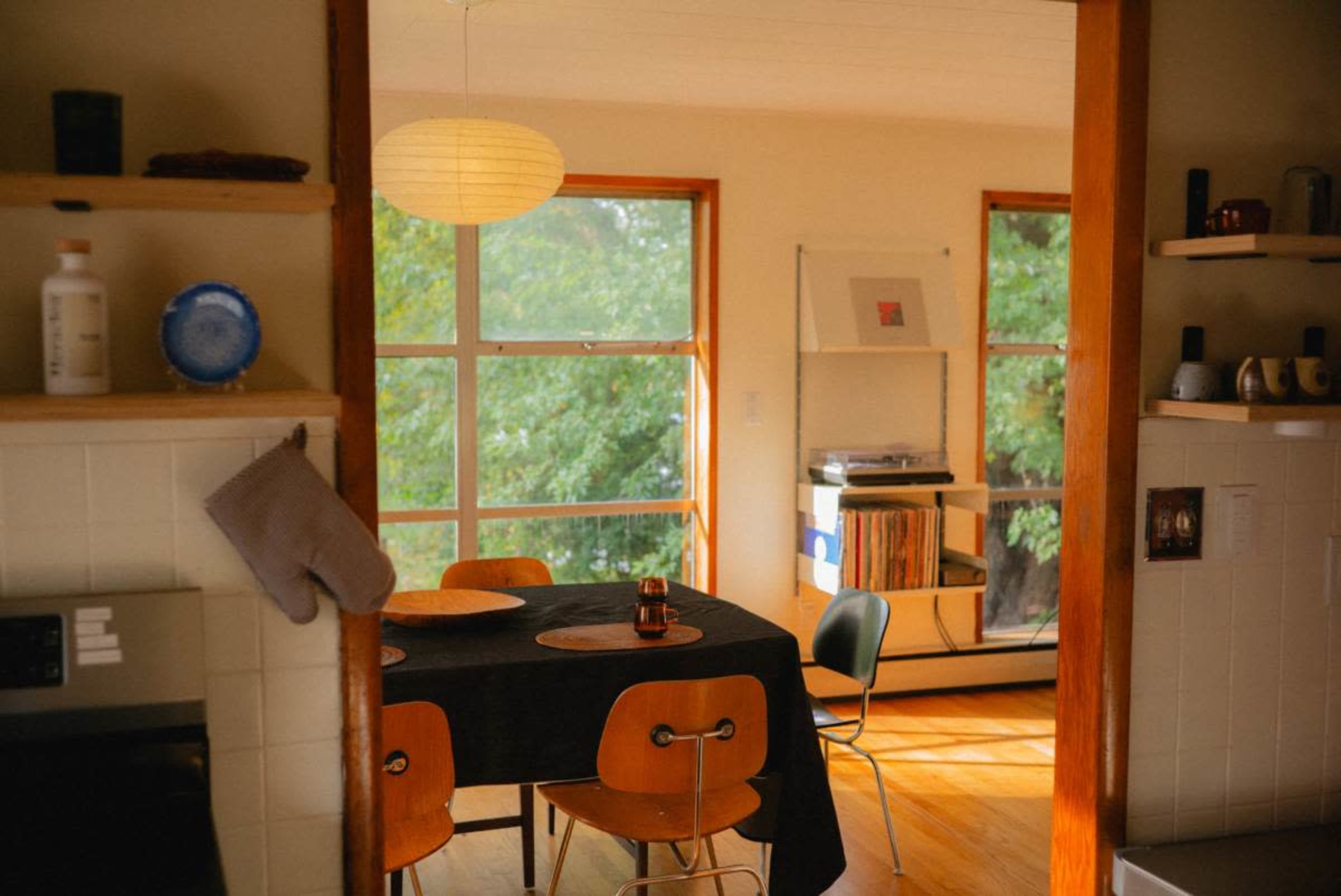 A kitchen and dining area features a table with four chairs, a large window with a view of greenery, and light streaming into the room.