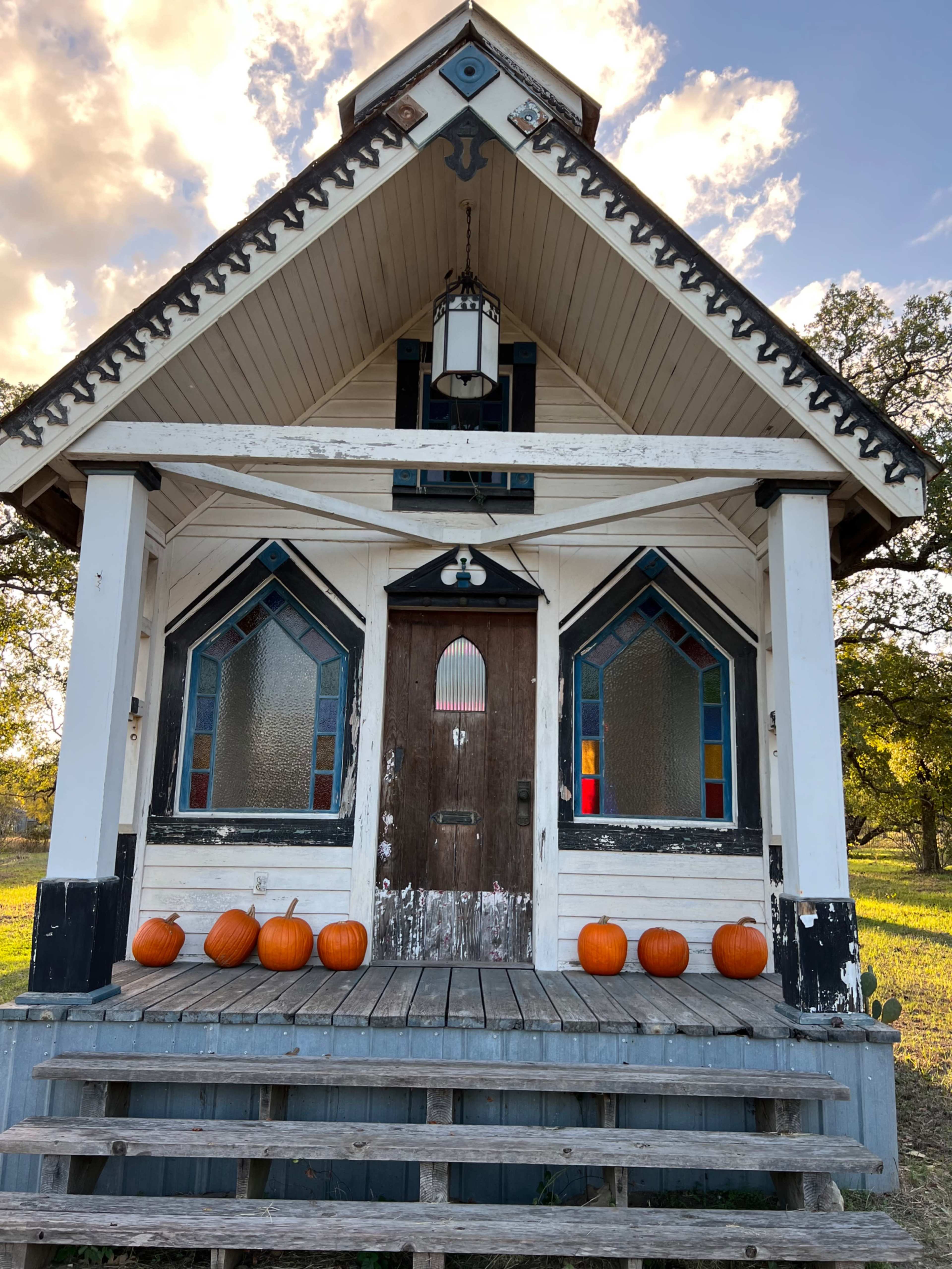 A small, decorative house with stained glass windows and pumpkins lined up on the front steps.