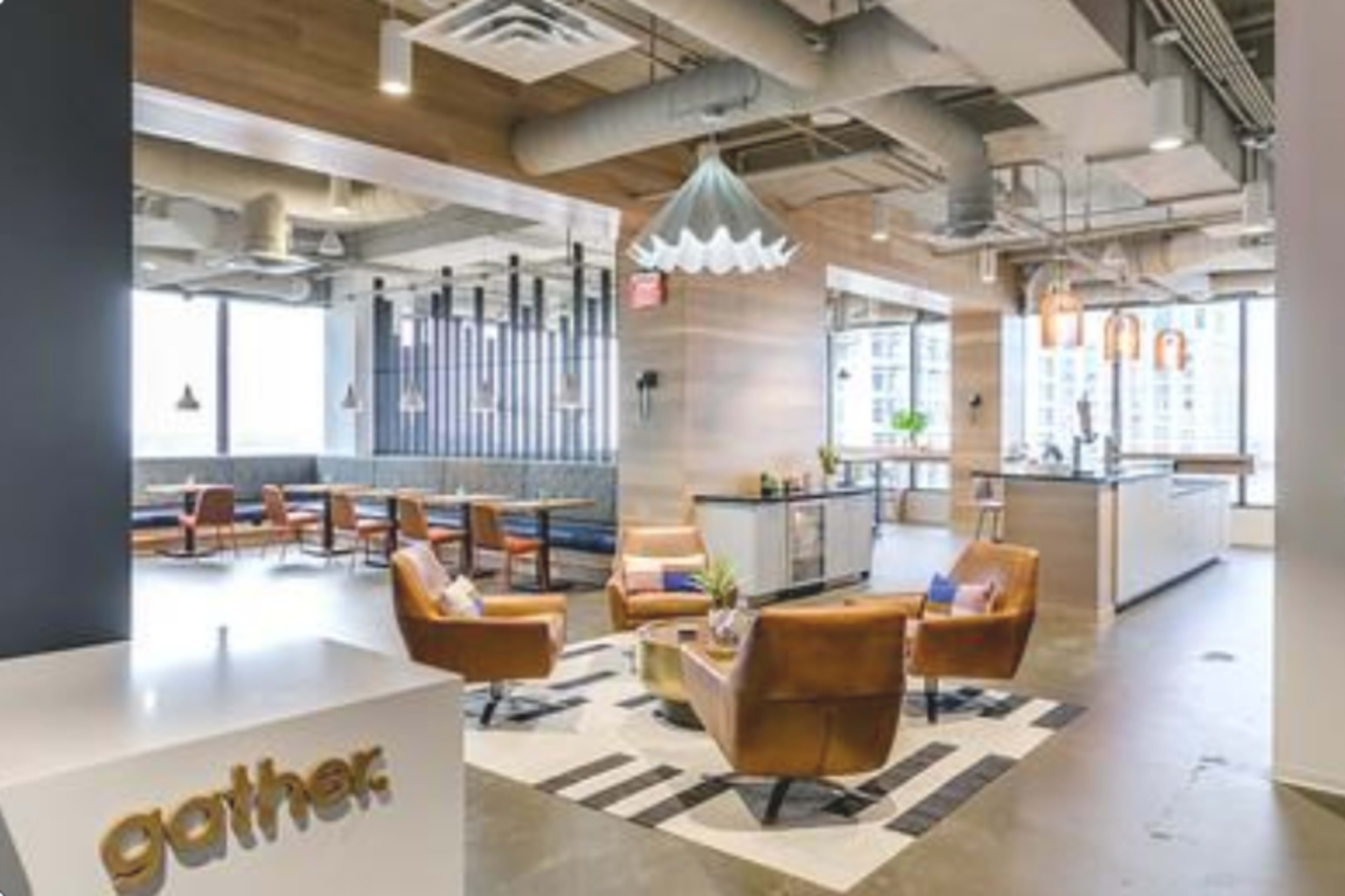 The image shows a modern office lounge area with brown leather chairs, a patterned rug, and large windows providing natural light.